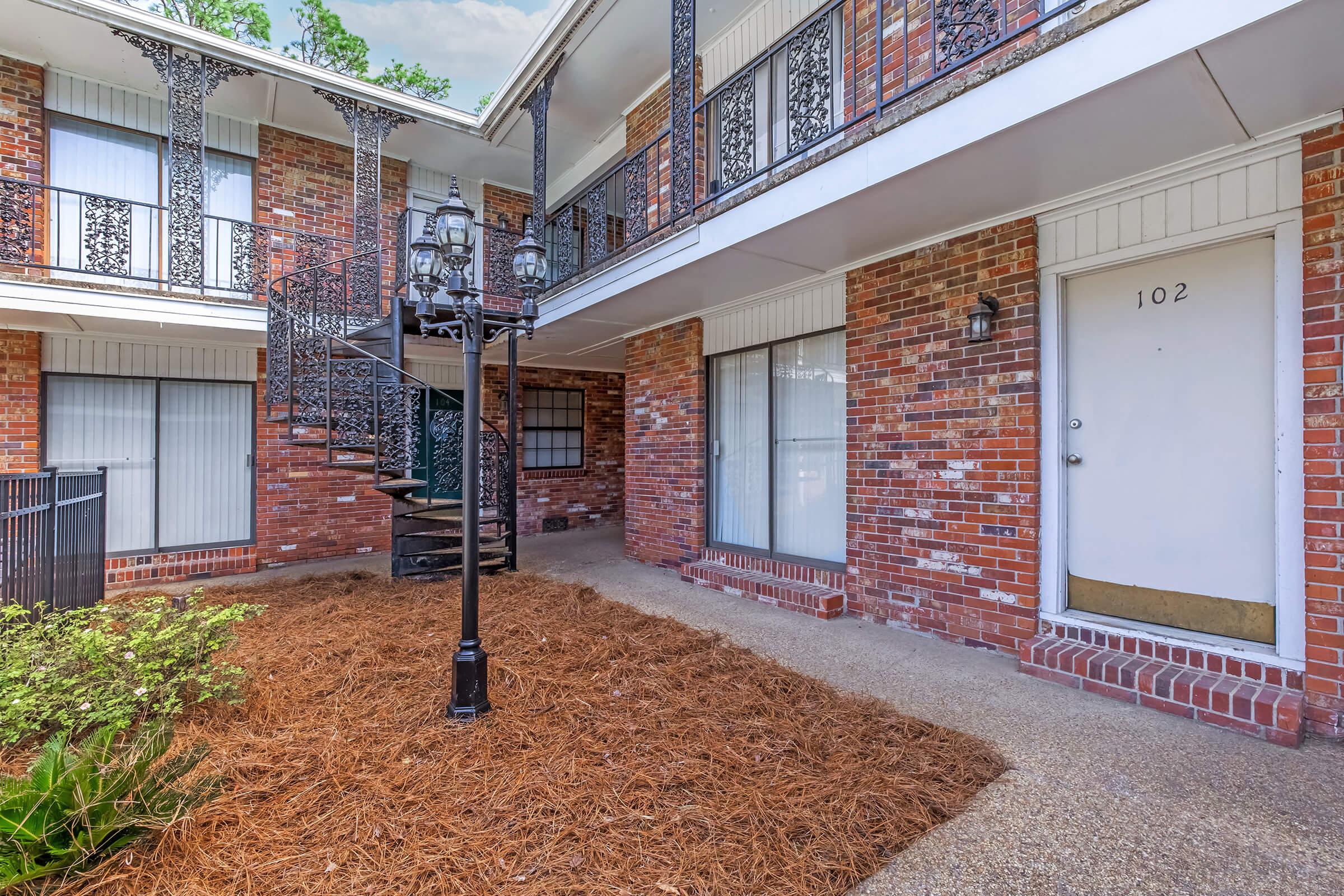A brick apartment building with a spiral staircase leading to upper levels. The courtyard features a lamp post and well-maintained landscaping with pine straw. Several windows are visible, showcasing the entrance to unit 102.