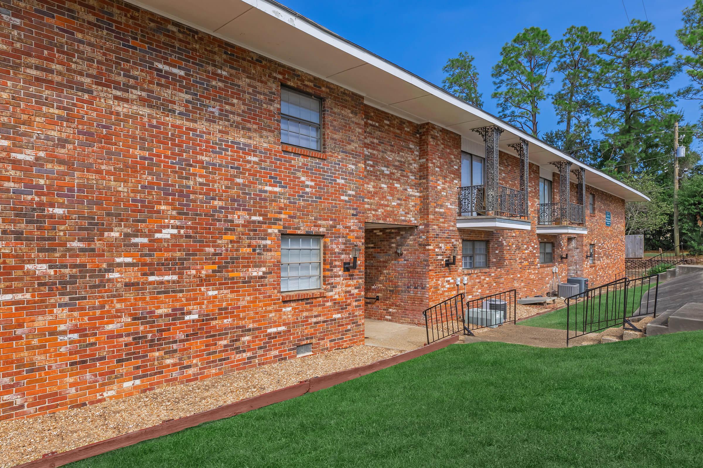 Exterior view of a brick apartment building with multiple units. The building features a red-brown brick facade, white window frames, and balconies with wrought iron railings. A grassy area is in front, and there are concrete stairs leading to the entrance. Tall trees and a clear blue sky are visible in the background.
