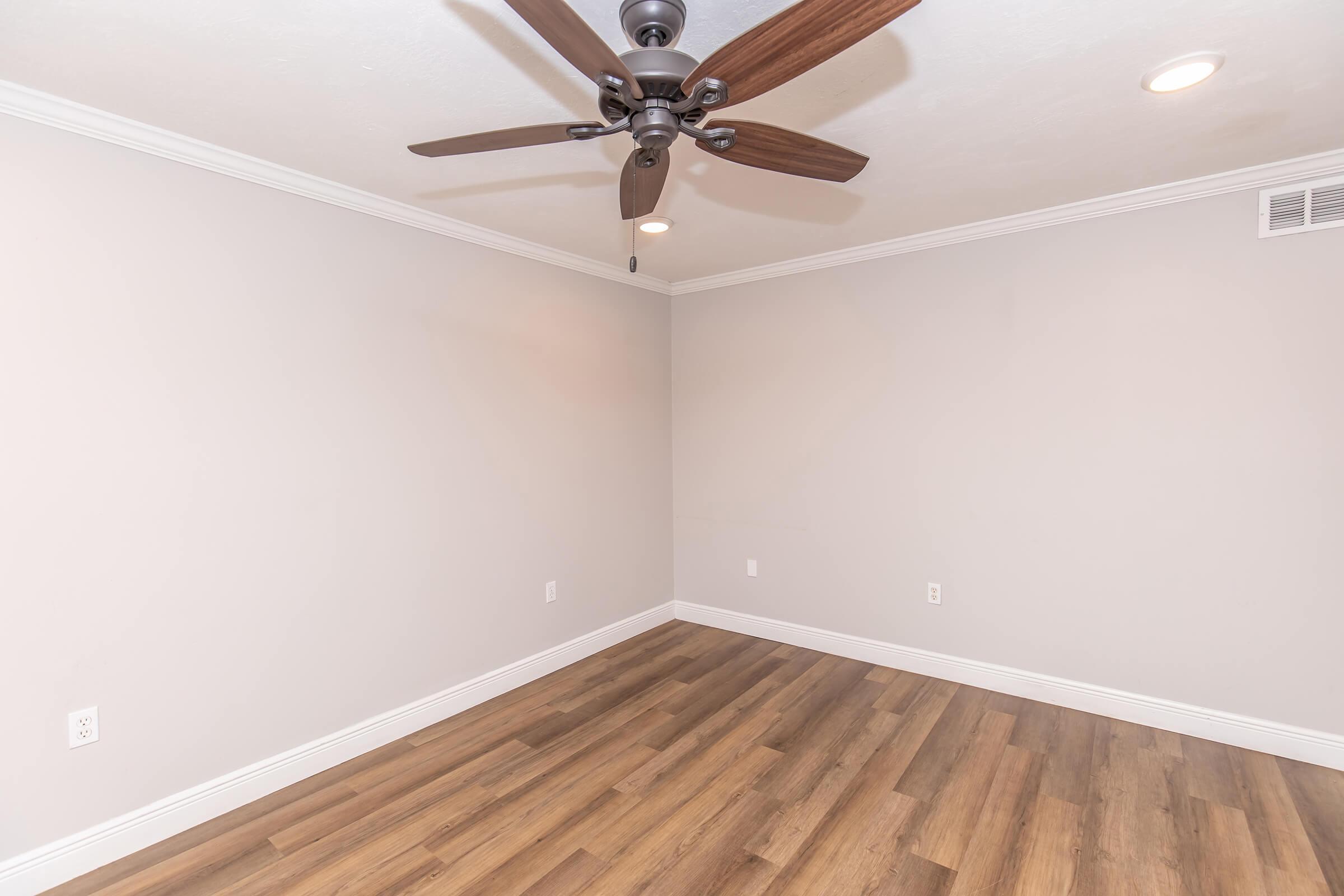 A spacious, empty room with light gray walls and light wood flooring. A ceiling fan with wooden blades is installed in the center, and there are recessed lights on the ceiling. The room features crown molding along the edges, creating a bright and open atmosphere.