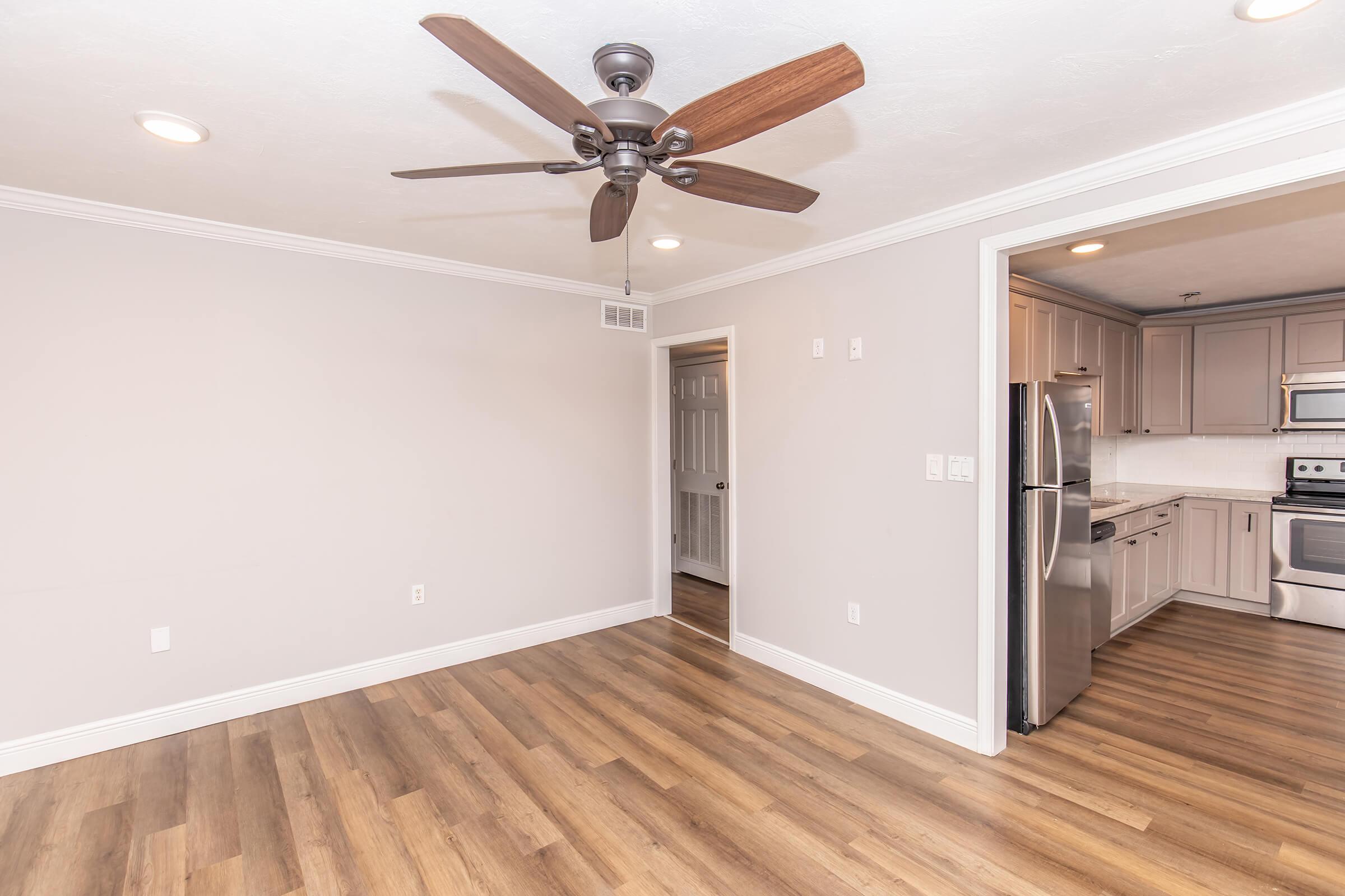 A spacious, modern living area featuring light gray walls, a ceiling fan with wooden blades, and warm wood-style flooring. To the right, an open kitchen with stainless steel appliances is visible, behind a doorway leading to another room. Natural light enhances the inviting atmosphere.