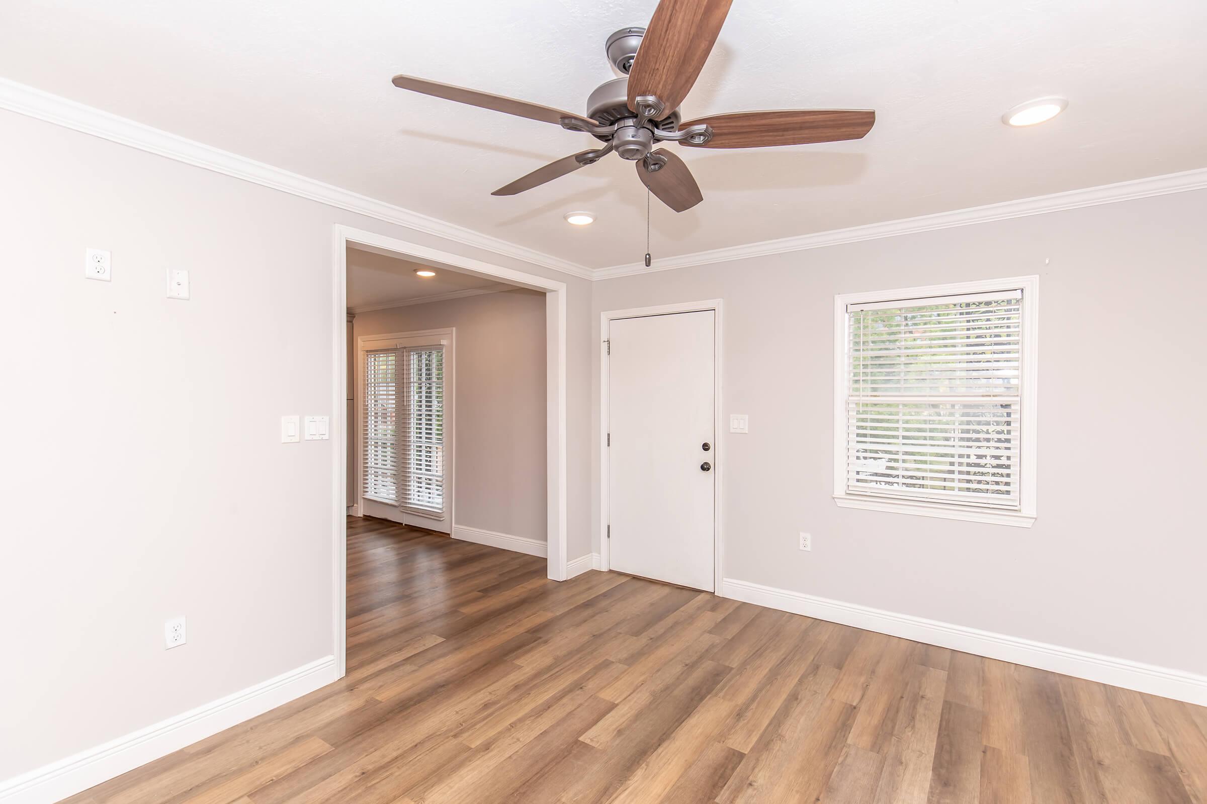 A bright, empty room with light gray walls and wooden flooring. A ceiling fan with wooden blades is visible. There's a doorway to another room and a closed white door. The window has plantation shutters, allowing natural light to fill the space.
