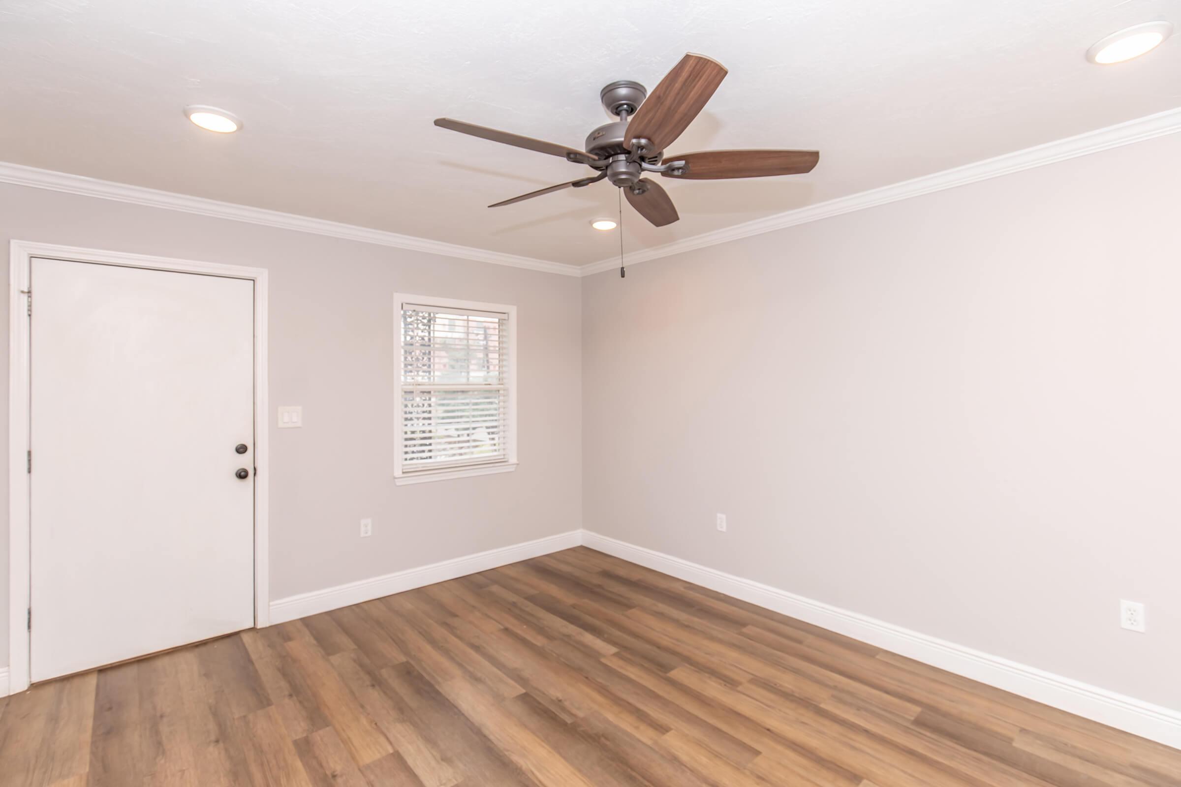 An empty room featuring light-colored walls, a wooden floor, a ceiling fan with brown blades, and a door on the left. A small window allows natural light in, and there are recessed lights in the ceiling, creating a bright and open atmosphere.