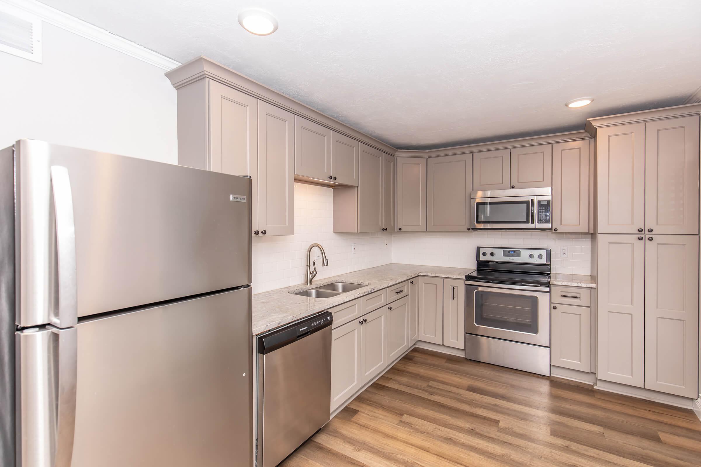 A modern kitchen featuring light gray cabinets, stainless steel appliances including a refrigerator, microwave, and oven. The countertop is a subtle stone texture, and there's a stainless steel sink with a faucet. The floor is made of wood, creating a warm ambiance.