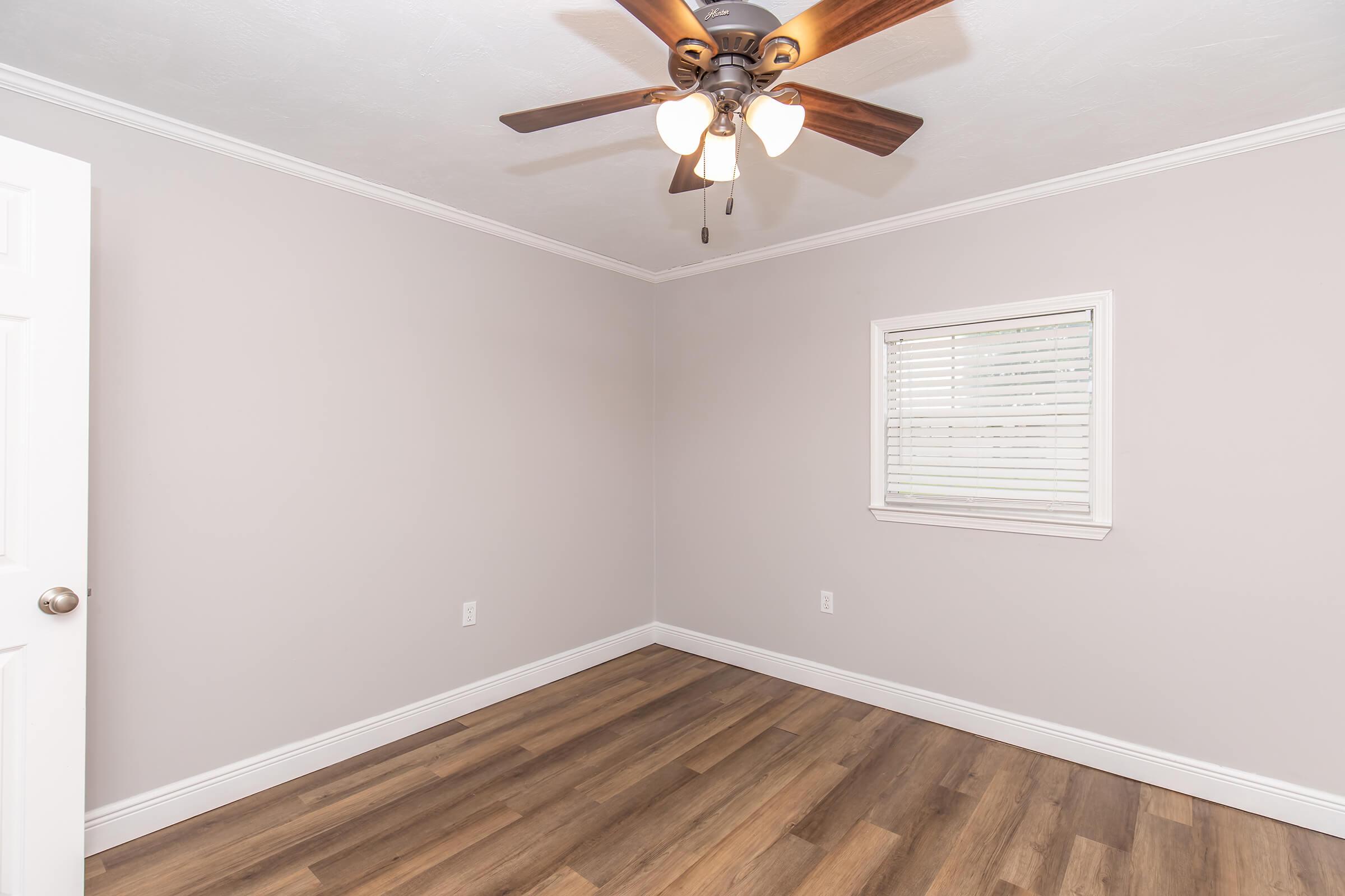 A simple, empty room with light gray walls and a wooden floor. There’s a white ceiling fan with three lights hanging from the ceiling. A small window with white blinds lets in natural light. The room has a clean, modern look, suitable for various uses.