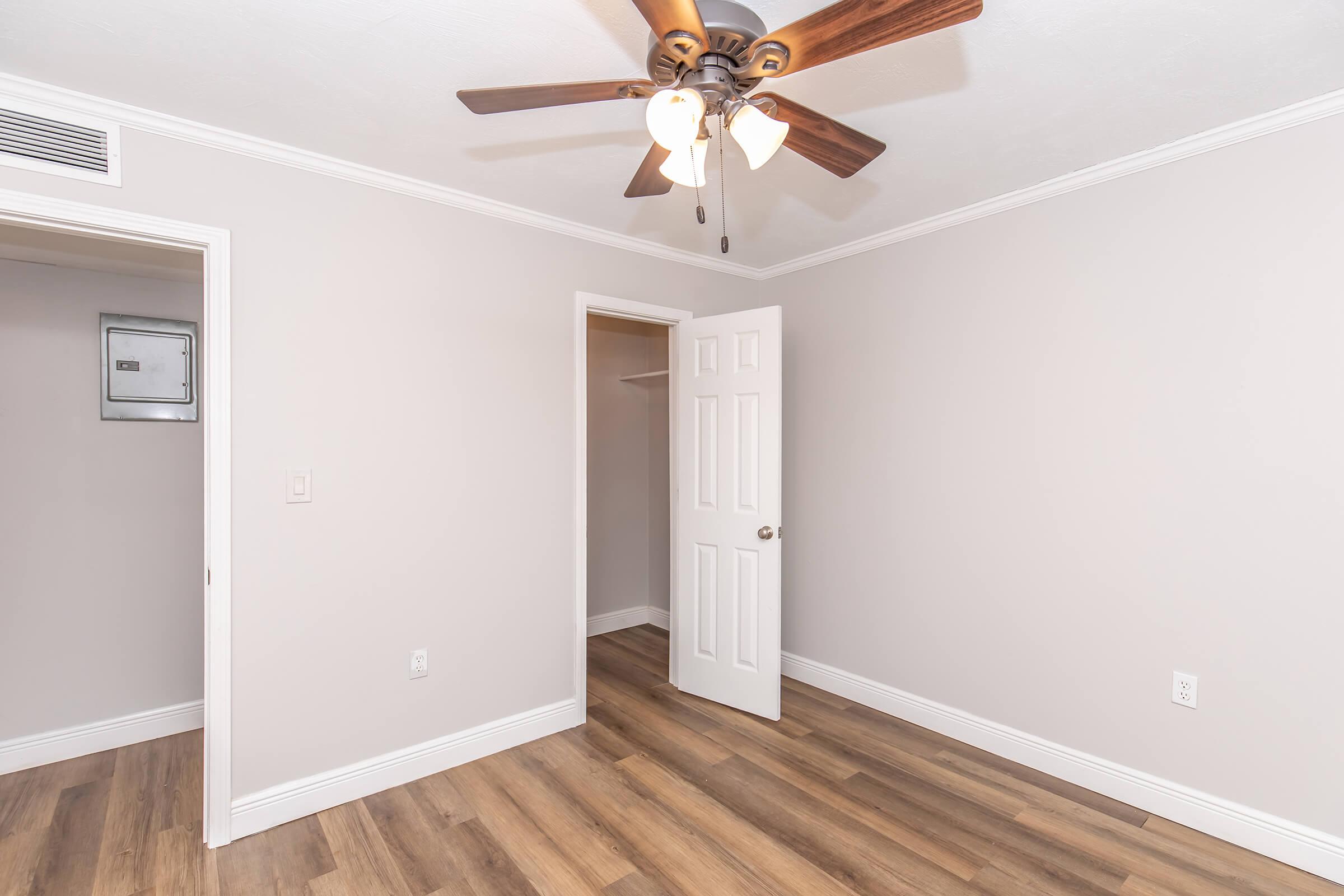 A well-lit room with light gray walls, a ceiling fan with four bulbs, and wood laminate flooring. One doorway leads to a closet and another doorway is slightly open, revealing an adjacent space. The room is tidy and minimally decorated, creating a spacious feel.