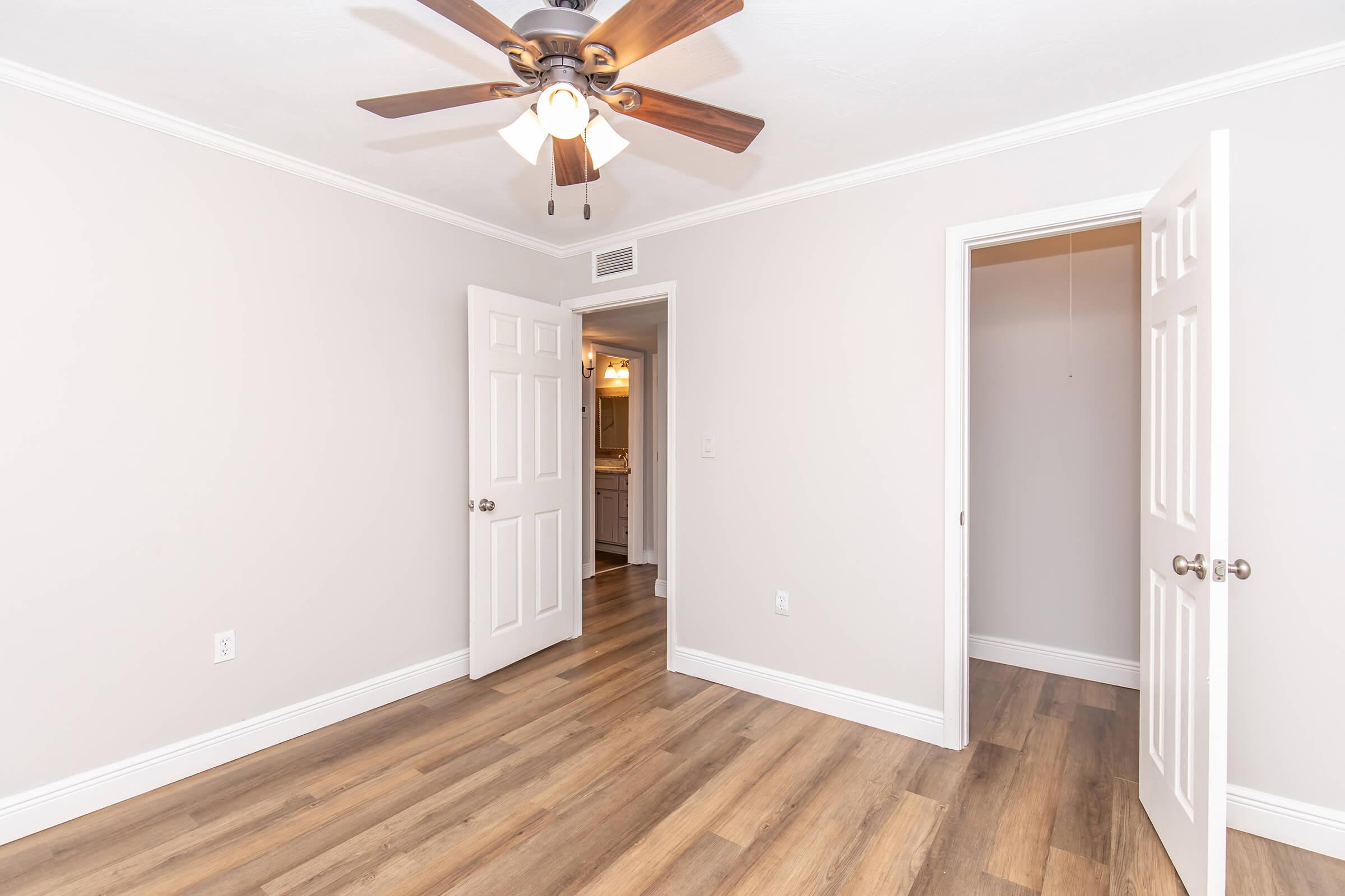 A well-lit, empty room featuring light-colored walls and hardwood flooring. There is a ceiling fan with wooden blades, and two white doors leading to other areas, one slightly open. The room has a clean, modern aesthetic suitable for various uses, such as a bedroom or office.
