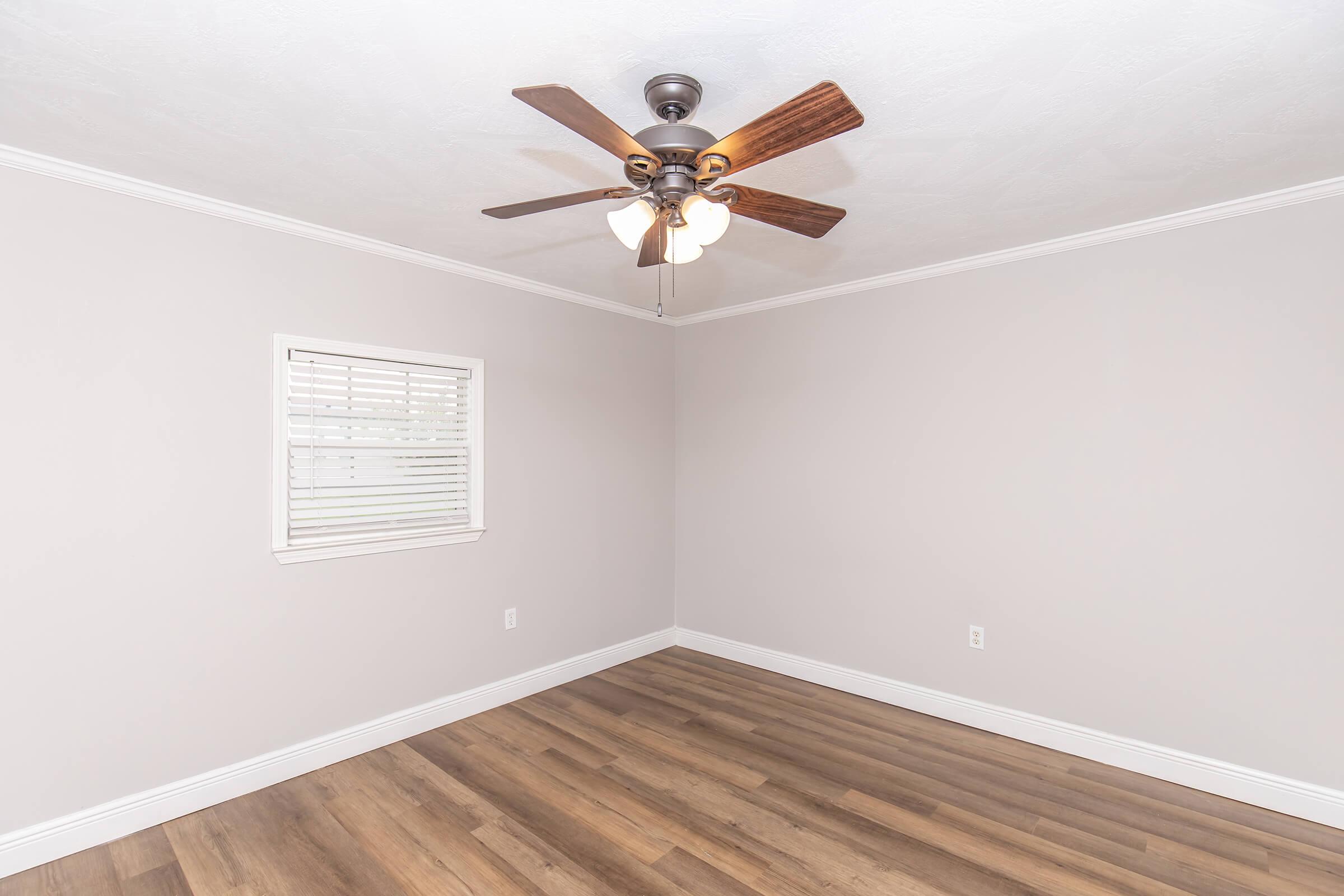 Empty room with light gray walls, a ceiling fan with three bulbs, and a window with white blinds. The floor is wooden, and there are no furniture or decorations visible. The space appears clean and ready for occupancy.