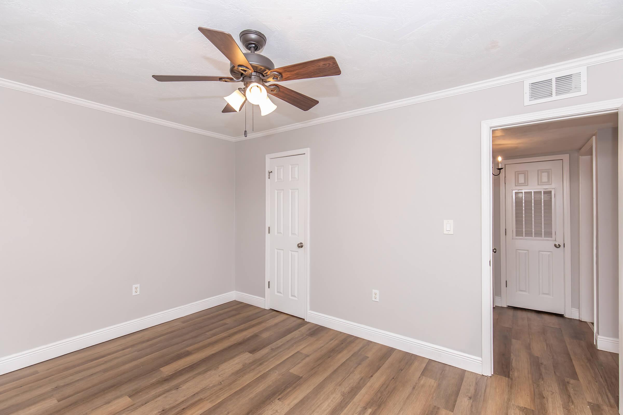 A lightly colored room featuring a ceiling fan with wooden blades, hardwood flooring, and white crown molding. One wall has a closed door, while another leads to a hallway with an additional door visible. The room has a bright and airy feel, suitable for various decor styles.