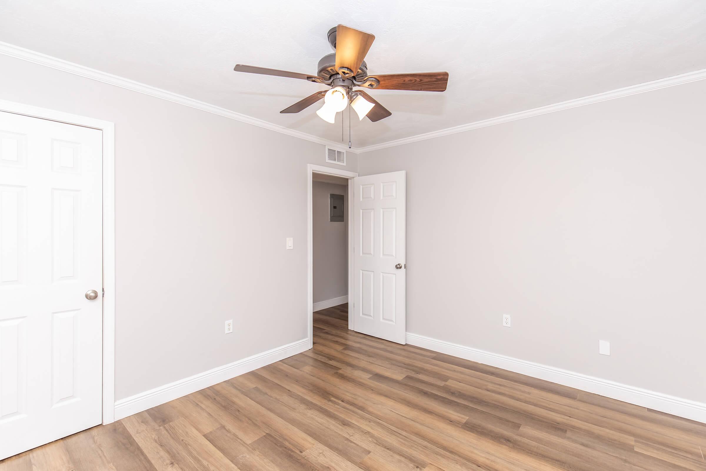 A well-lit room with light gray walls and a ceiling fan featuring wooden blades. It has hardwood flooring and two white doors, one closed and one slightly ajar, leading to other areas. The room has a clean and minimalistic aesthetic, suitable for a bedroom or office space.