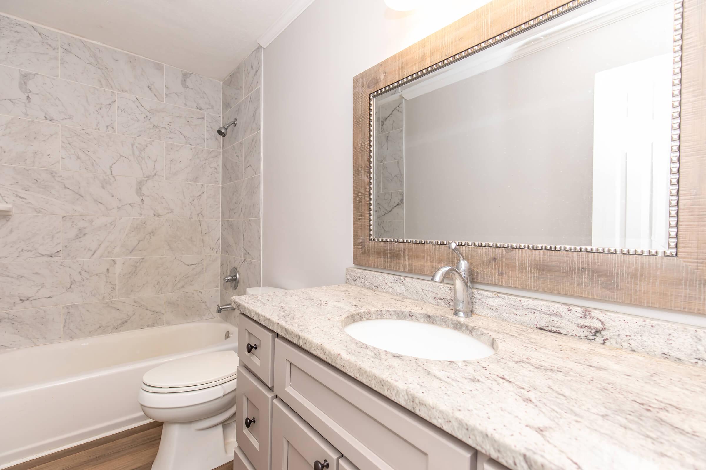 A modern bathroom featuring a marble-tiled wall, a white bathtub with a shower fixture, and a stylish countertop with a circular sink. The vanity has gray cabinetry and a large framed mirror above it, with soft lighting enhancing the serene atmosphere.