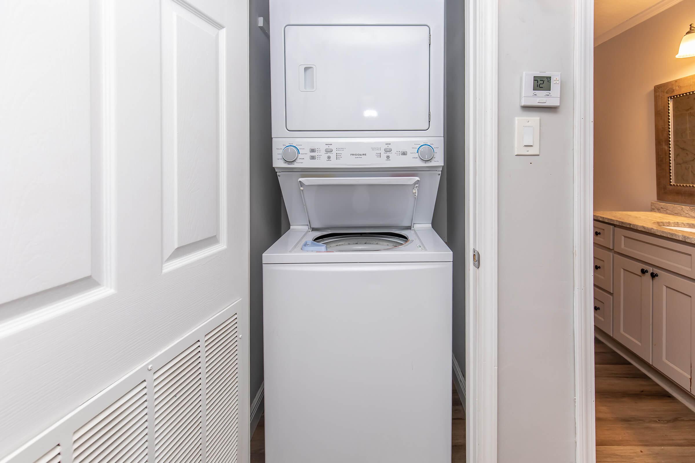A compact white stacked washer and dryer unit situated between a wall and a closed door. The dryer is on top, with a control panel featuring blue knobs and buttons. The surrounding walls are light-colored, and there is a thermostat on the wall nearby.