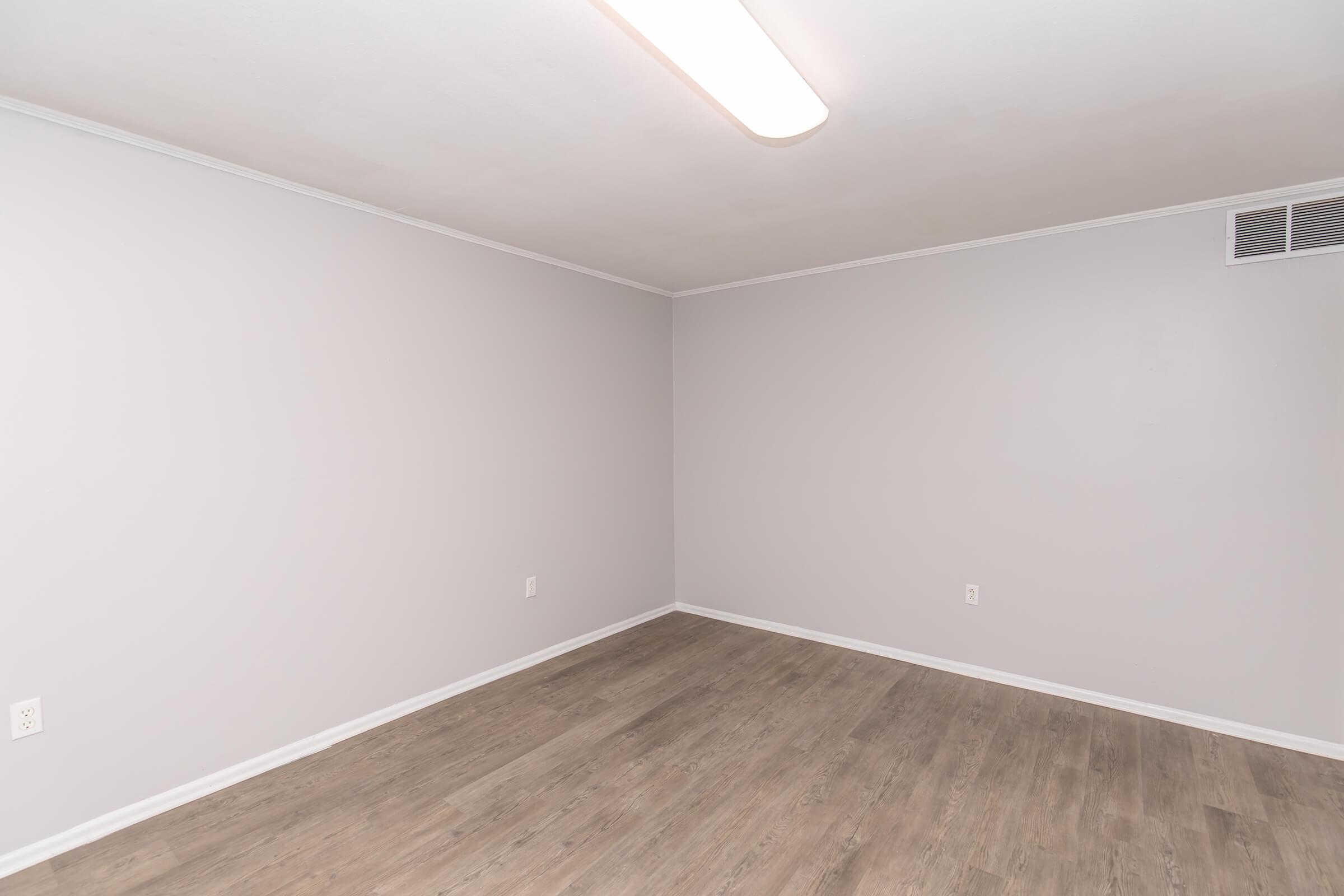 Empty room with light gray walls and a wooden floor. A ceiling light fixture provides illumination. There are no furnishings or decorations, creating a spacious and minimalistic appearance. A wall vent is visible, but no other features are present.