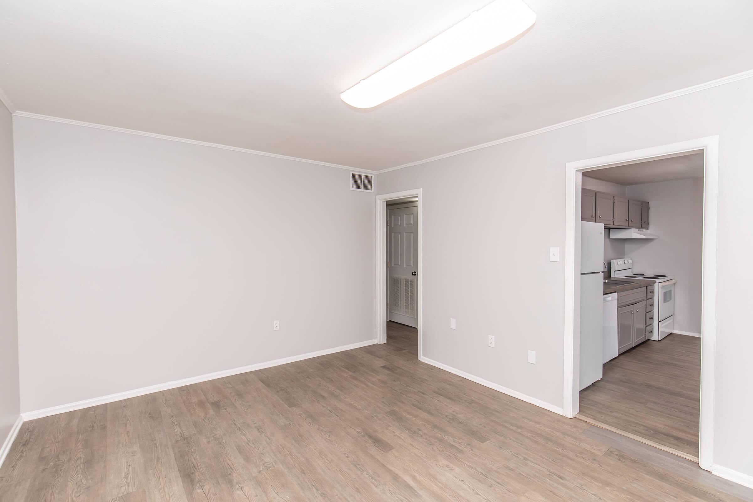 A well-lit living area with light gray walls and a hardwood floor, featuring an open layout. In the background, there is a doorway leading to a kitchen with white appliances and cabinetry. The space appears clean, empty, and ready for furnishings.