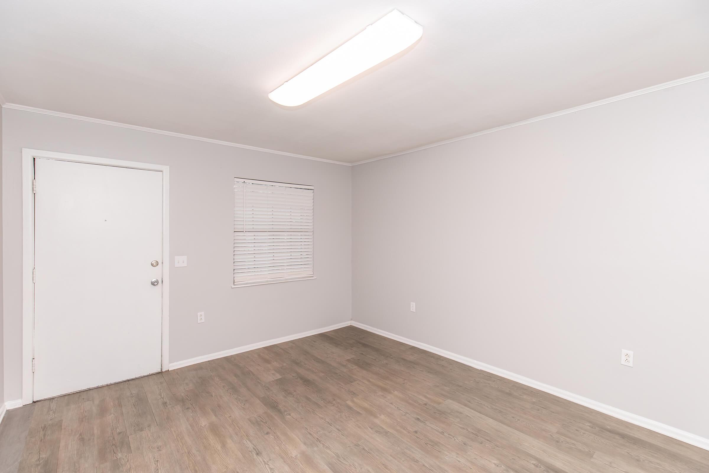 Empty room with light beige walls and a light wood floor. There is a closed white door on the left, a small window with white blinds, and a ceiling light fixture. The space is unfurnished and has no visible decorations, creating a minimalist and clean appearance.