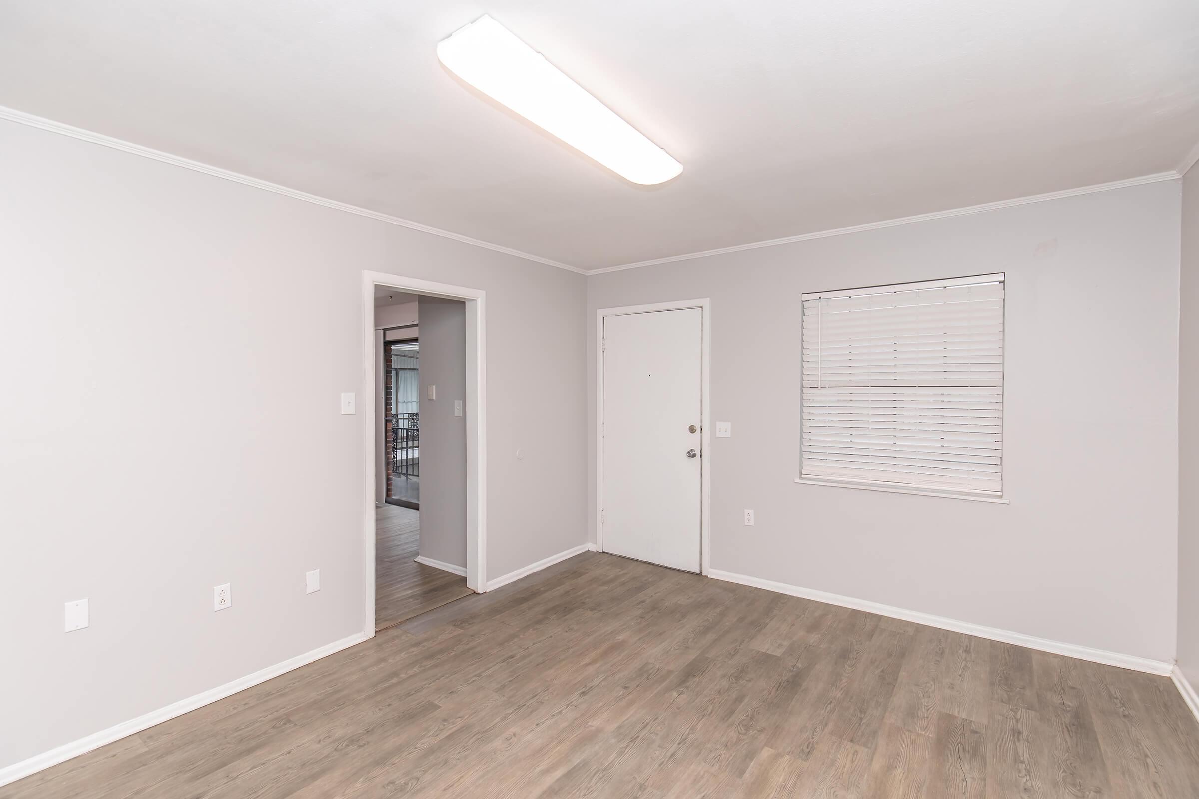 Empty room with light-colored walls and a wooden floor. A ceiling light illuminates the space. There's a closed door on the left leading to another room, and a window with blinds on the right. The overall atmosphere is minimalistic and inviting.