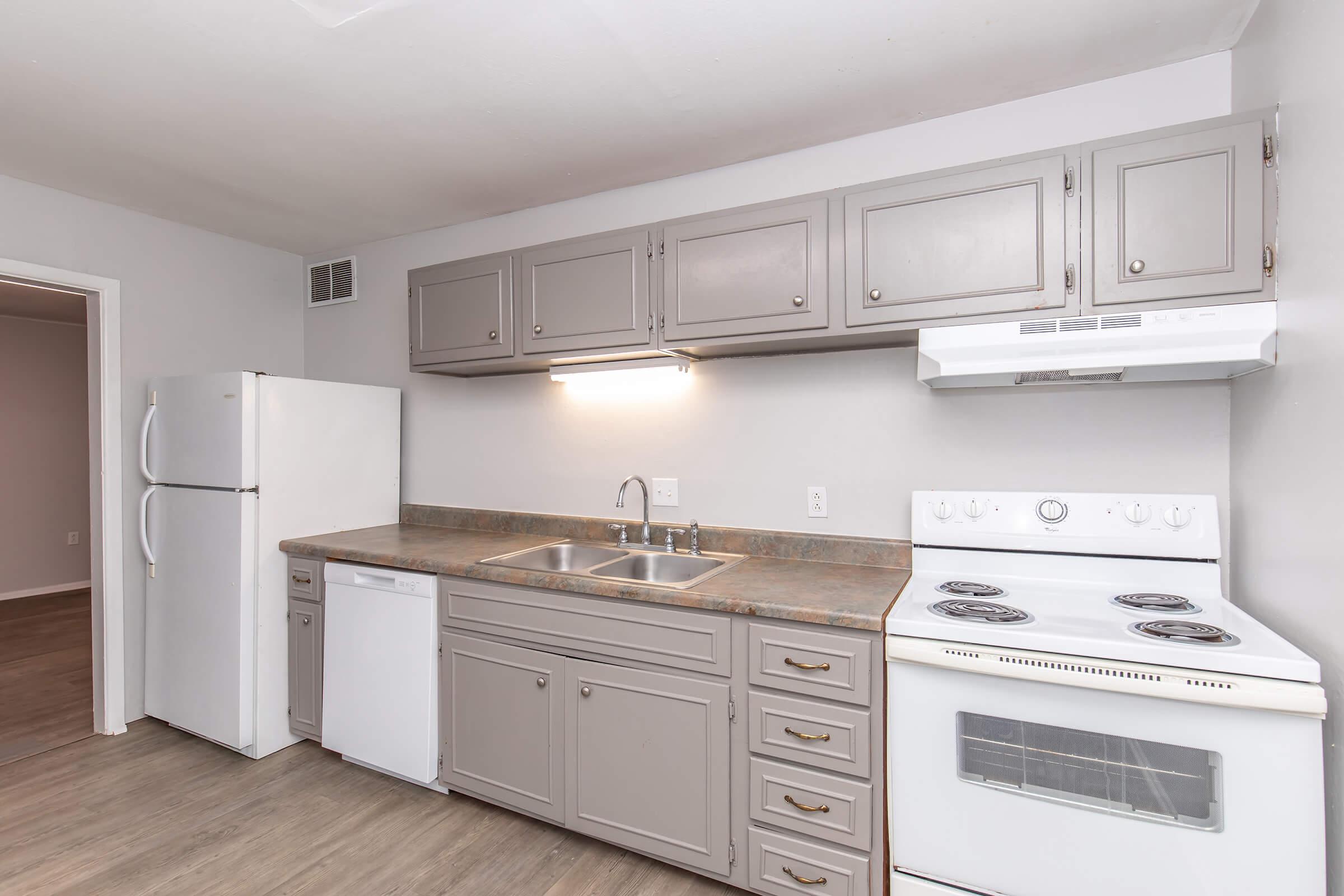 A modern kitchen featuring gray cabinets, a white refrigerator, a dishwasher, and a stove with an oven. The countertop is stone, and there is a double sink under a light fixture. The walls are painted a neutral color, and the flooring is a light wood laminate.