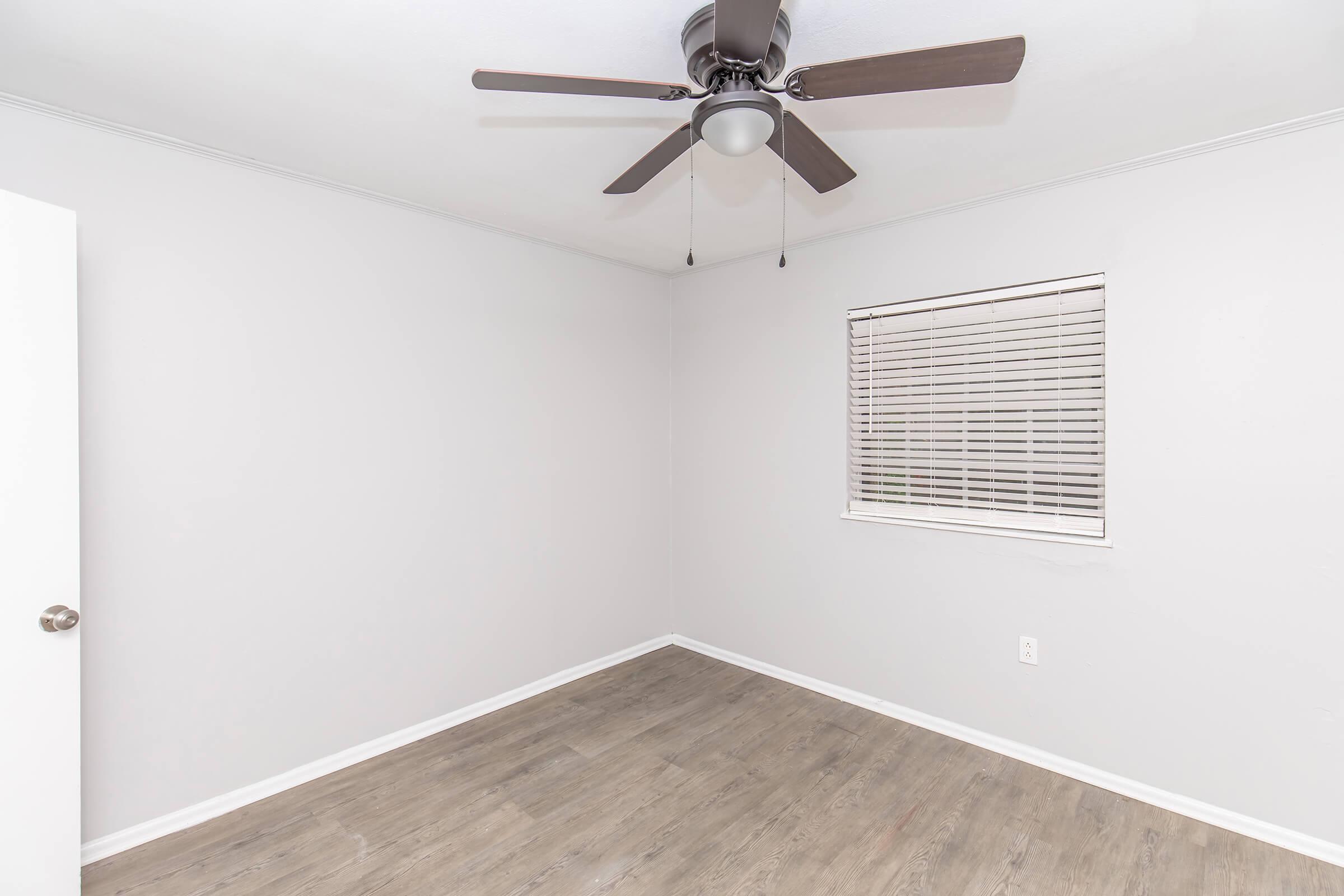 A clean, empty room with light gray walls and a wooden floor. A ceiling fan with dark wooden blades is installed. There is a white door on the left and a window with white blinds on the right, allowing natural light to enter the space. The room appears spacious and well-maintained.