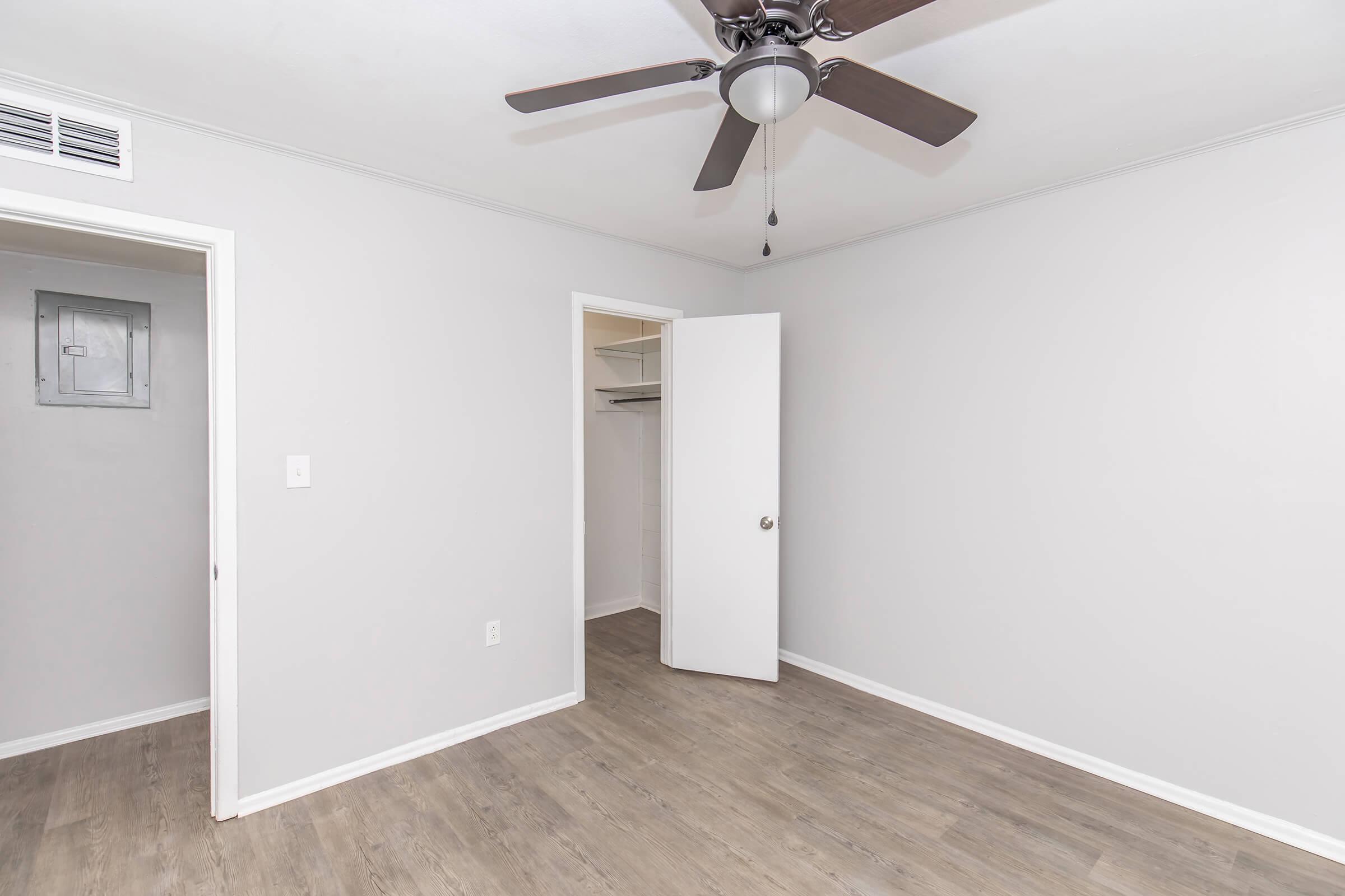Empty room with light gray walls and a ceiling fan. One door is partially open, leading to a closet, while another door is visible on the left. The flooring is a light wood, and there are no furnishings in the space.