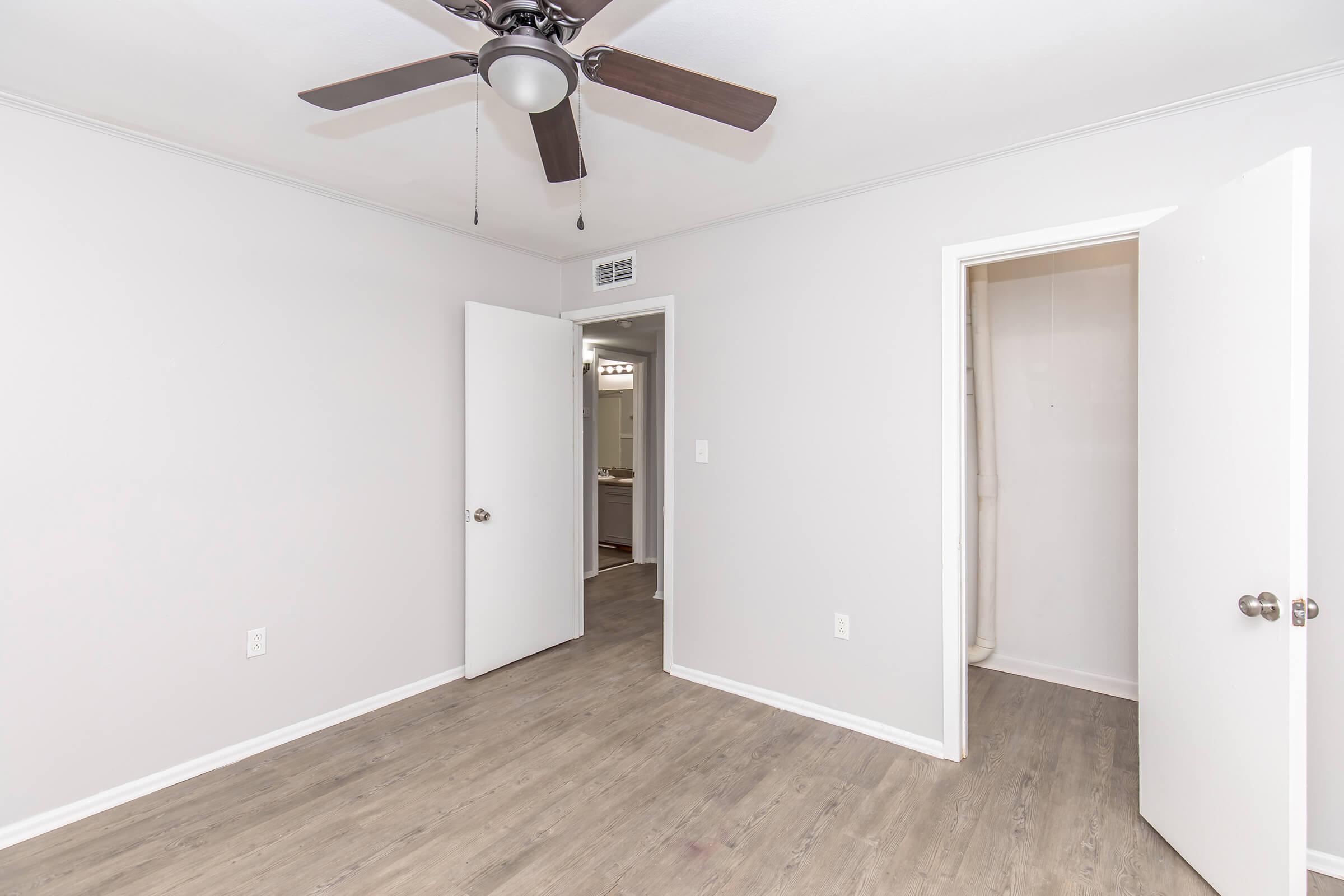 A simple, unadorned room with light gray walls and wood-like flooring. There are two doors: one leads to a bathroom, and the other is slightly ajar, showing a closet space. A ceiling fan is mounted in the center, adding to the room's minimalistic design.