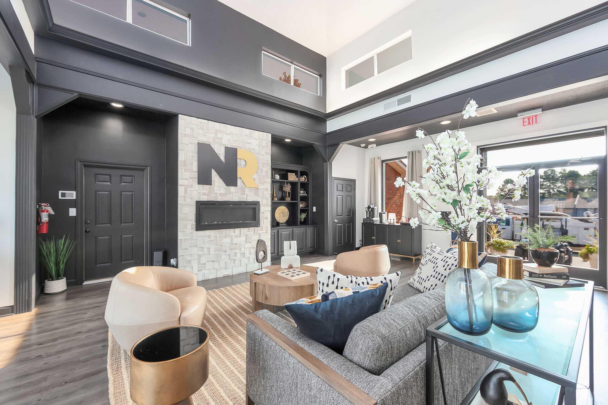 A modern living room featuring stylish furniture, including a gray couch, beige armchairs, and decorative vases. The walls are primarily black with a textured white accent wall displaying the letters "NR." Large windows let in natural light, showcasing a cozy, contemporary atmosphere.