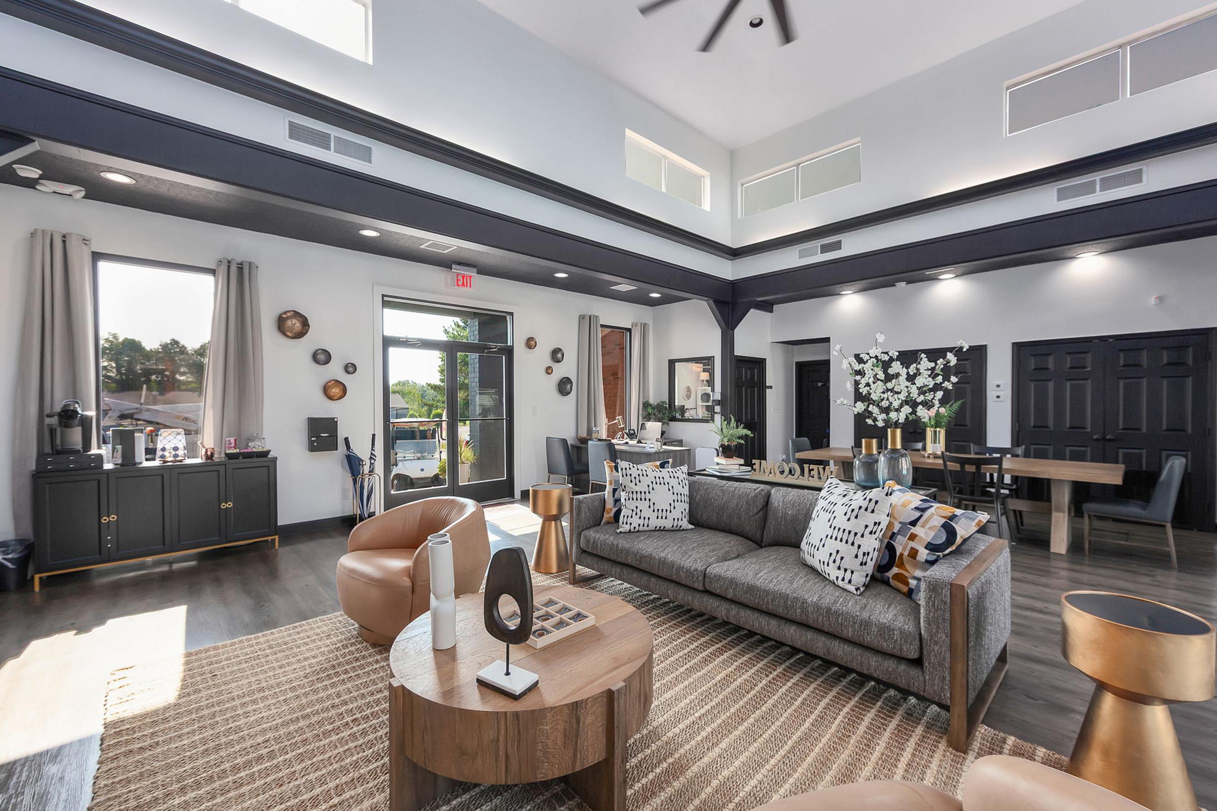 A modern living room featuring a gray sofa adorned with decorative pillows, a round coffee table, and stylish armchairs. The space has large windows that allow natural light, a high ceiling with a fan, and elegant decor including plants and wall art. The flooring is wood-like, adding warmth to the contemporary design.
