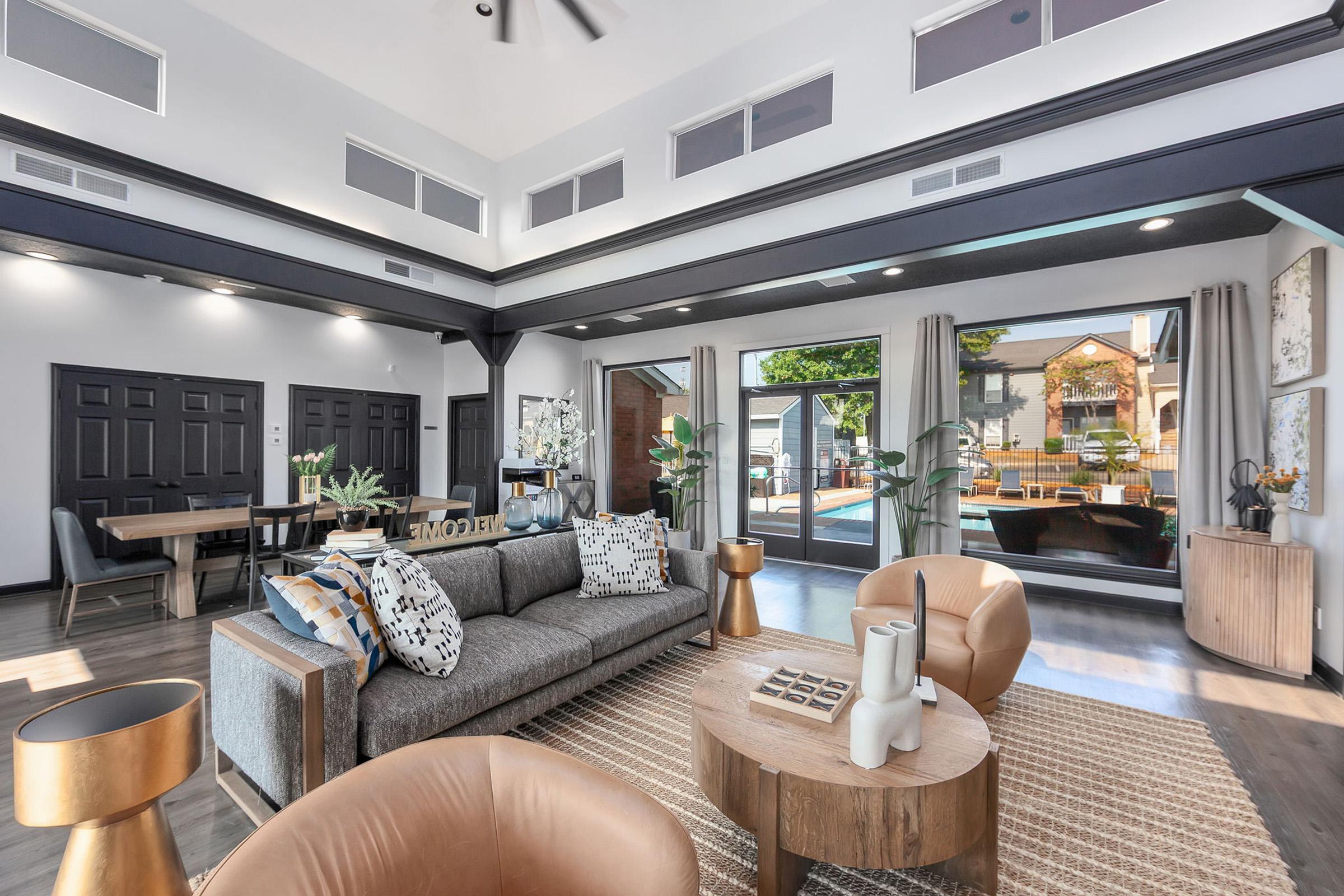 A modern, stylish living room featuring a gray sofa with decorative pillows, two tan armchairs, a round wooden coffee table, and a textured rug. Large windows offer a view of an outdoor area, and the space is illuminated by natural light. Wood accents and sleek decor create a welcoming atmosphere.