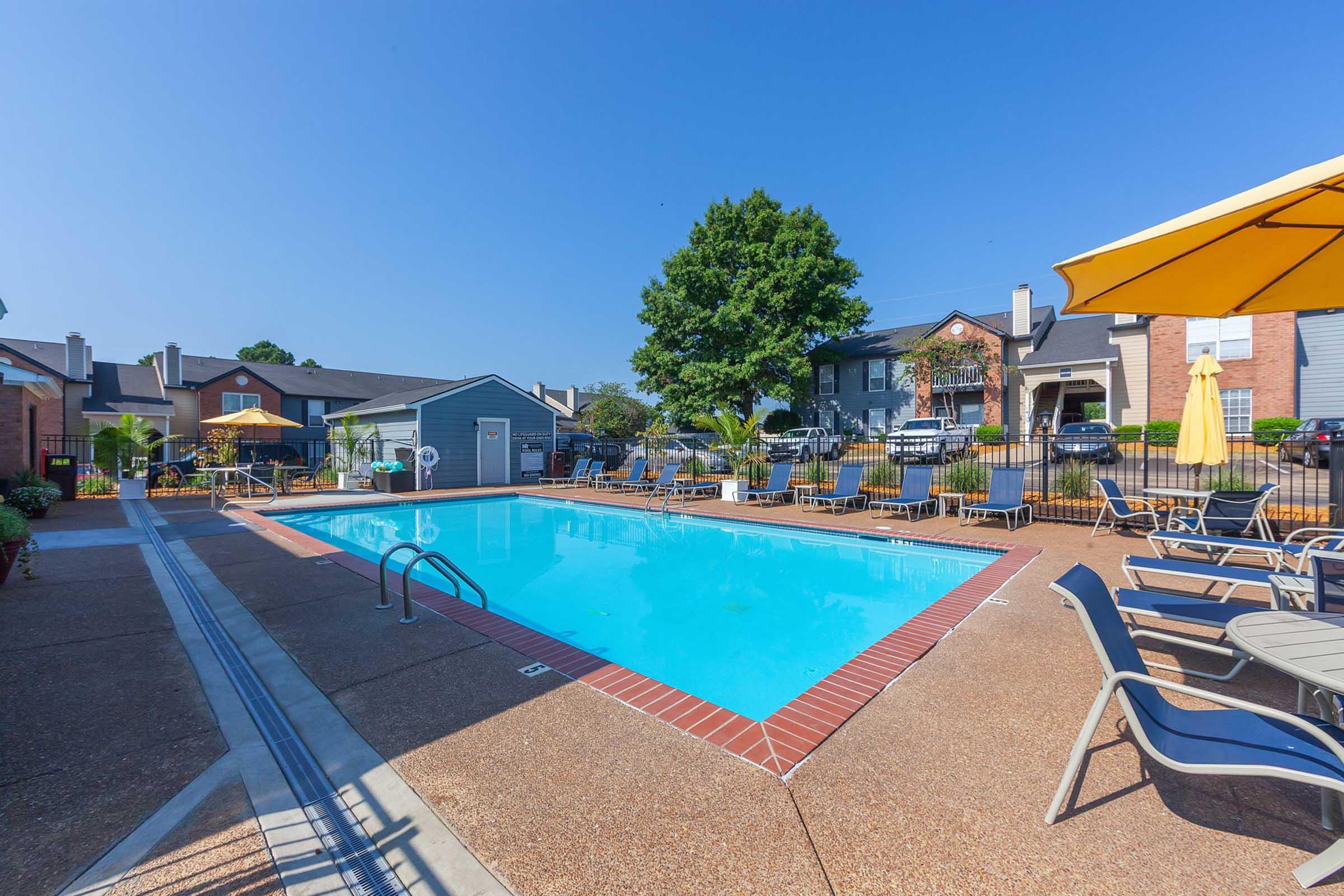 A clear blue swimming pool surrounded by lounge chairs and umbrellas, located in a residential area. Green trees and buildings are visible in the background under a sunny blue sky. The pool area is well-maintained and inviting.