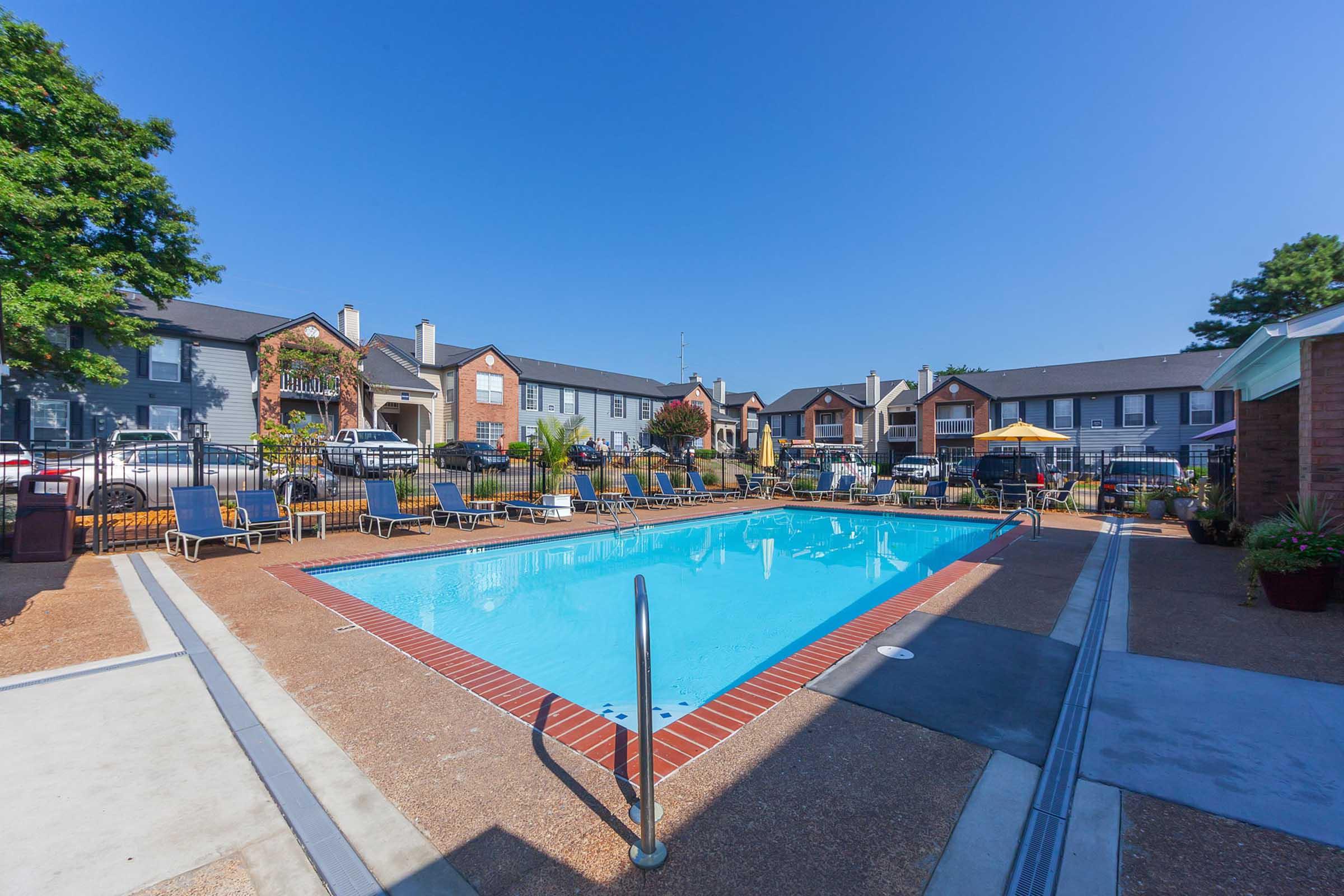 A clear blue swimming pool surrounded by lounge chairs, with apartment buildings in the background. Sunshine brightens the scene, creating a welcoming atmosphere with well-maintained landscaping and a few parked cars visible.