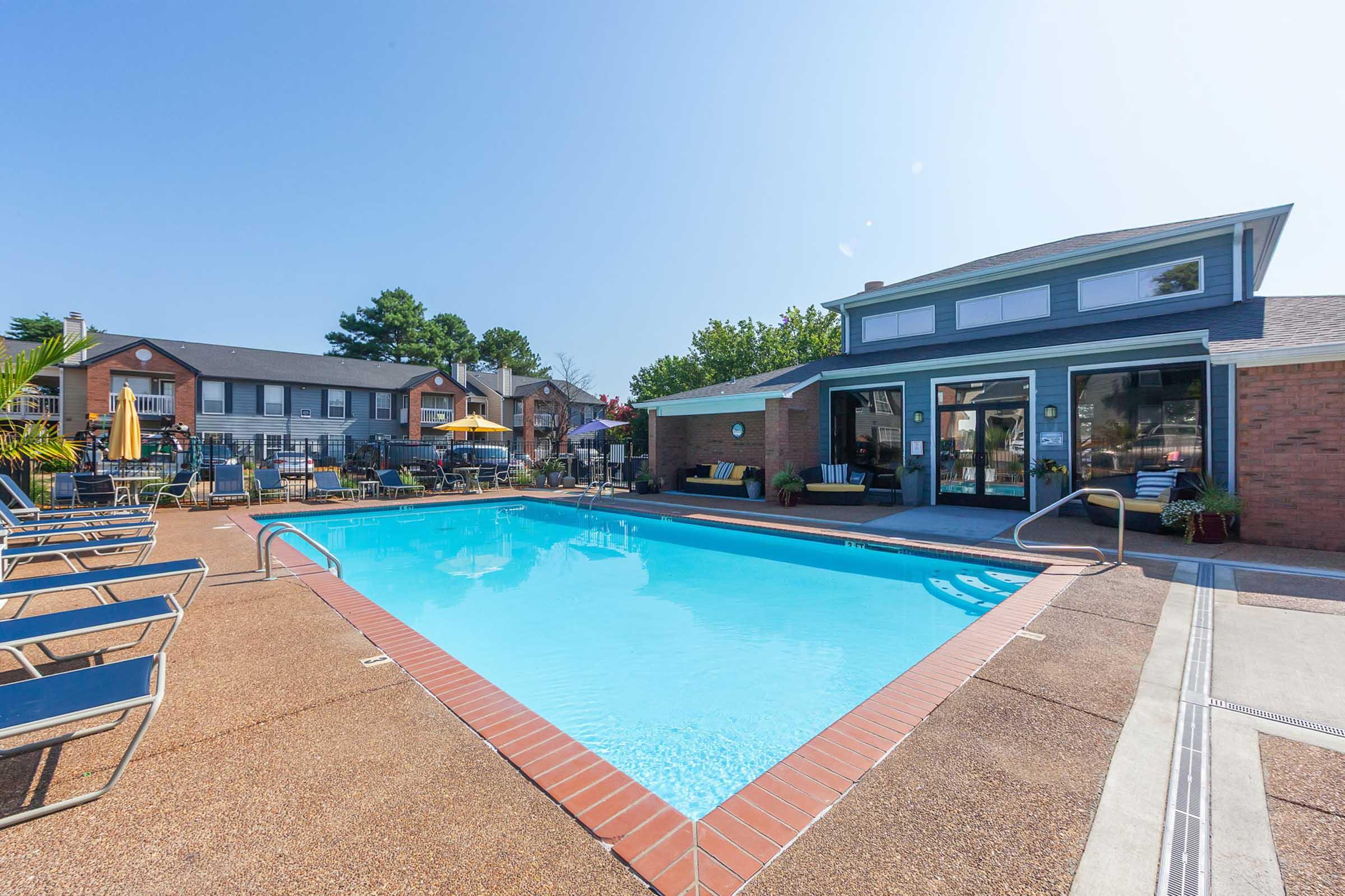 A crystal-clear swimming pool surrounded by lounge chairs, with a brick building in the background that features large windows. Sunlight shines brightly overhead, and patio furniture is visible around the pool area, suggesting a relaxed outdoor atmosphere. Green trees and landscaping can be seen nearby.