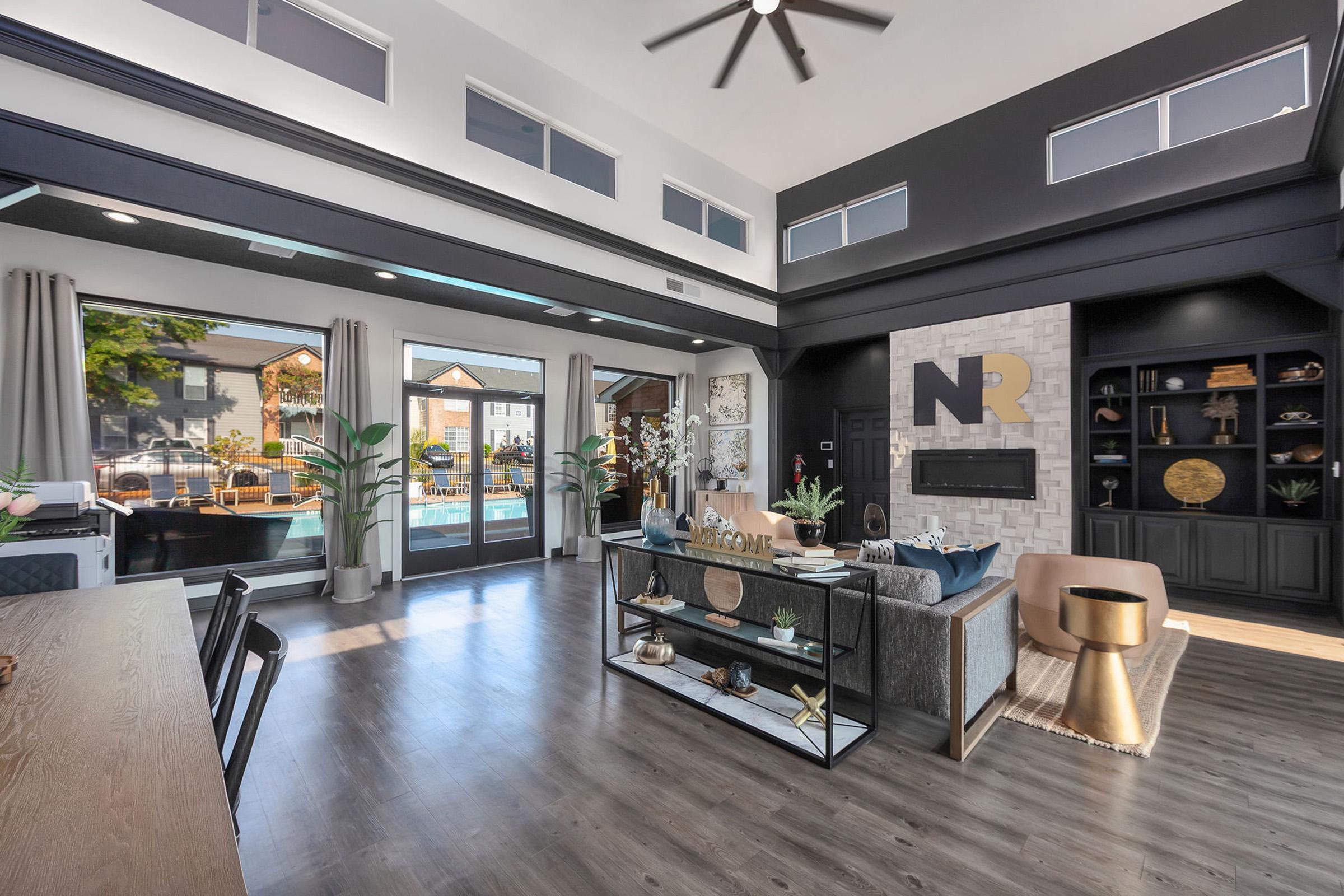 A modern living room featuring high ceilings, large windows, and stylish decor. The space includes a comfortable seating area with a coffee table, a decorative shelf, and potted plants. Natural light fills the room, enhancing the contemporary design elements and color scheme.