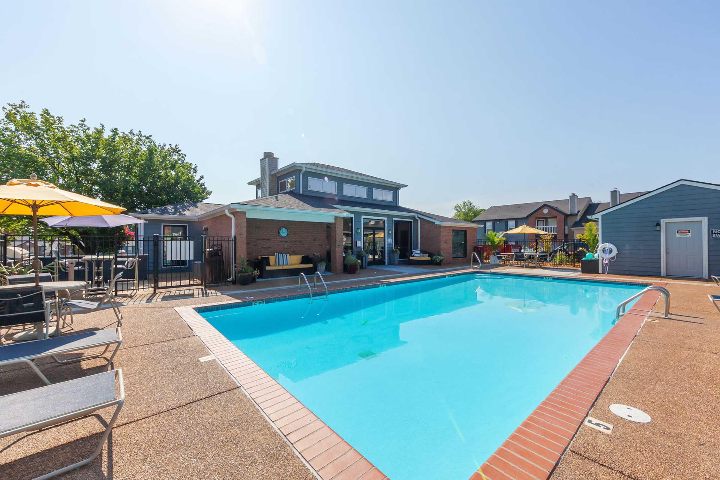 A clear blue swimming pool surrounded by a tan deck, lounge chairs, and umbrellas. In the background, a two-story building with large windows is visible, along with additional outdoor seating. Lush green trees provide shade, and the sky is bright and sunny.