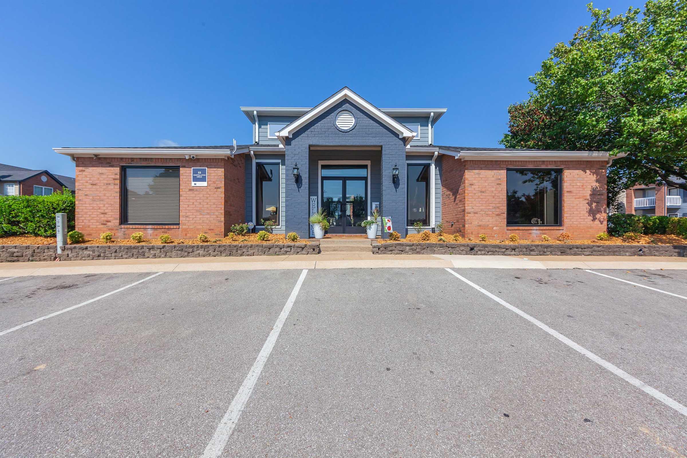 A modern building with a brick and dark gray facade, featuring large windows and a central entrance. The exterior includes landscaping with green plants and flower beds, parked cars in the foreground, and a clear blue sky.
