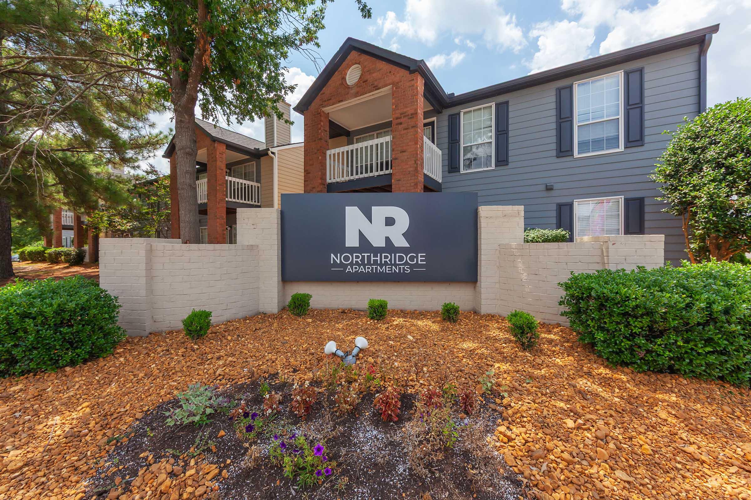 Sign for Northridge Apartments displayed at the entrance, featuring the letters "NR" prominently. The building has a modern design with brick and siding, surrounded by landscaped areas that include shrubs and flower beds. Cloudy sky in the background enhances the welcoming atmosphere.
