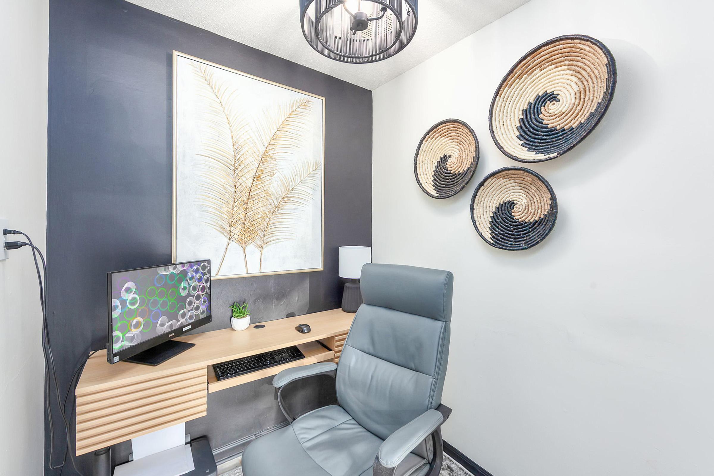 A cozy home office space featuring a wooden desk with a computer monitor, a chair, and a small potted plant. The walls are decorated with a large art piece of a feather and three woven wall hangings. Soft lighting from a pendant lamp creates a warm atmosphere.