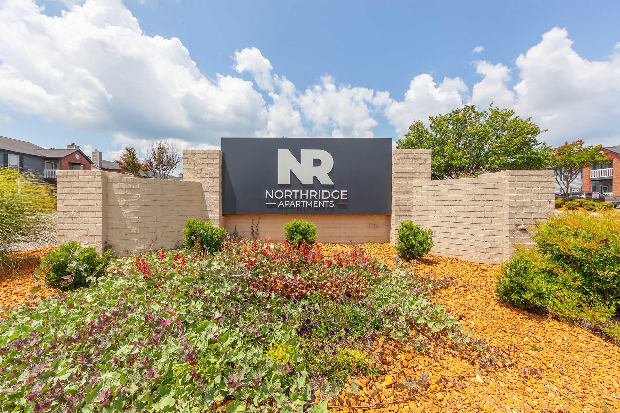 Sign for Northridge Apartments featuring the letters "NR" prominently displayed. The sign is surrounded by landscaped greenery and colorful flowers, with a clear blue sky and fluffy clouds in the background. The area has stone features and a well-maintained pathway, emphasizing a welcoming environment.