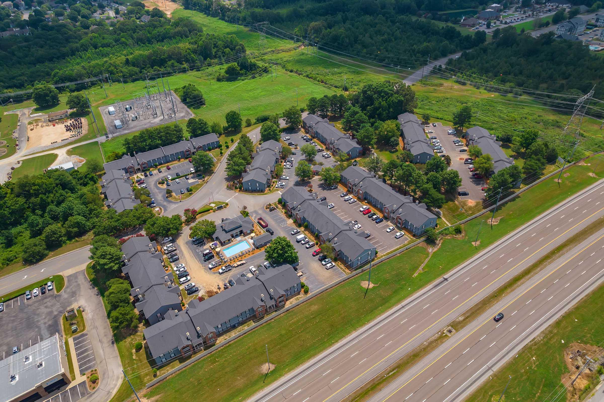 Aerial view of a residential complex featuring several two-story buildings arranged in a circular layout, surrounded by green lawns and trees. Adjacent to the complex is a busy highway with parked cars along the pathways, and power lines in the distance.