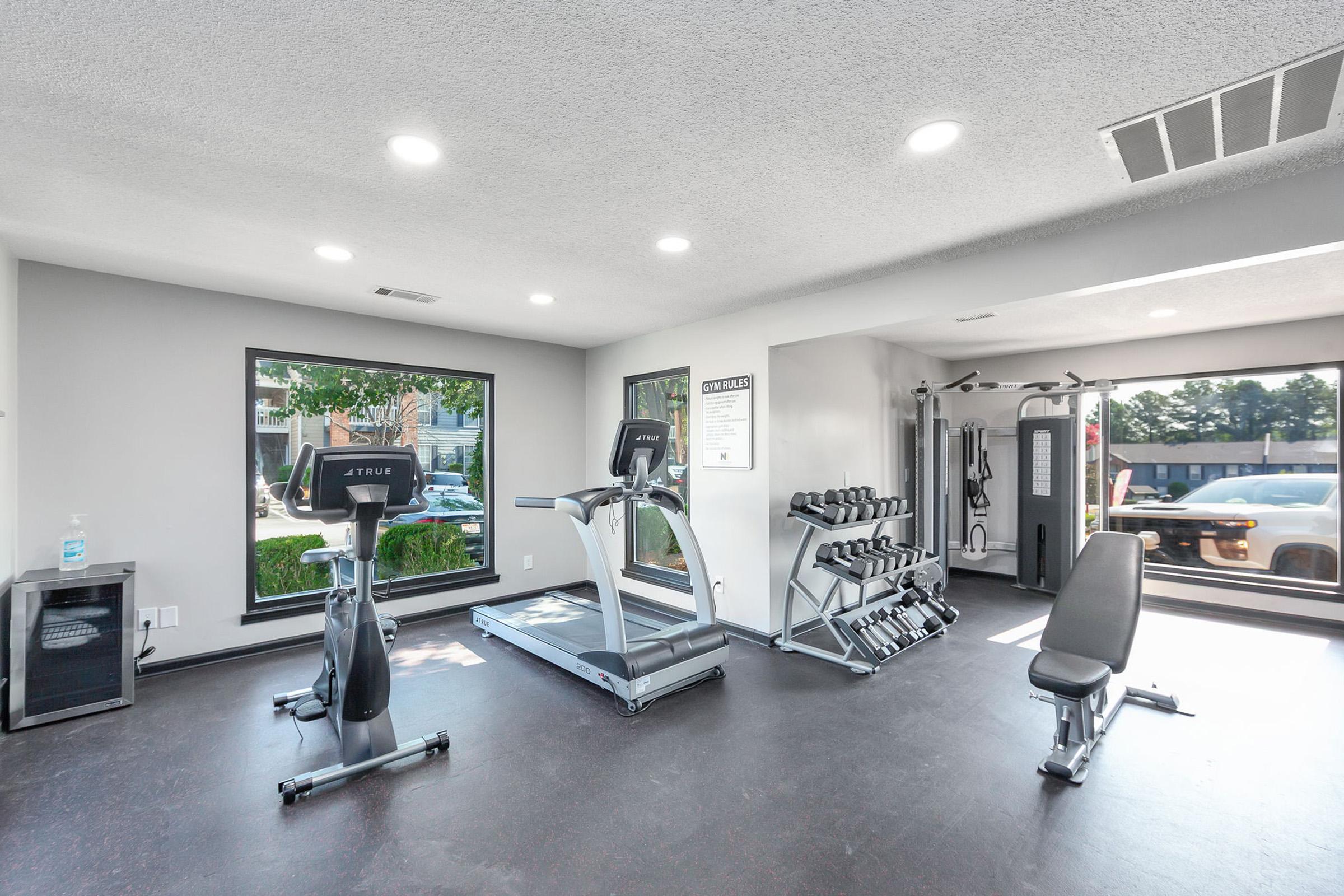 A modern gym interior featuring cardio equipment, strength training machines, and free weights. The space includes large windows allowing natural light, with a clean and minimalistic design. Nearby, a bottle of sanitizer is visible for hygiene.