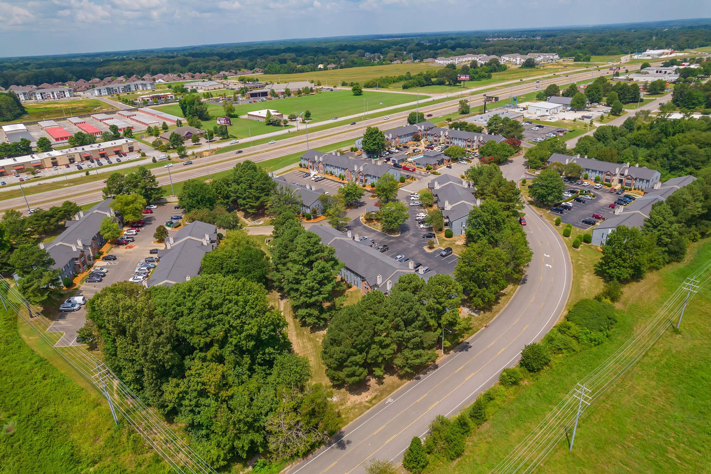Aerial view of a suburban area featuring a collection of low-rise buildings, parking lots, and tree-lined streets. Nearby, a major highway runs parallel to the development, with additional commercial properties visible in the distance. Lush green fields and trees surround the area, indicating a mix of residential and commercial use.