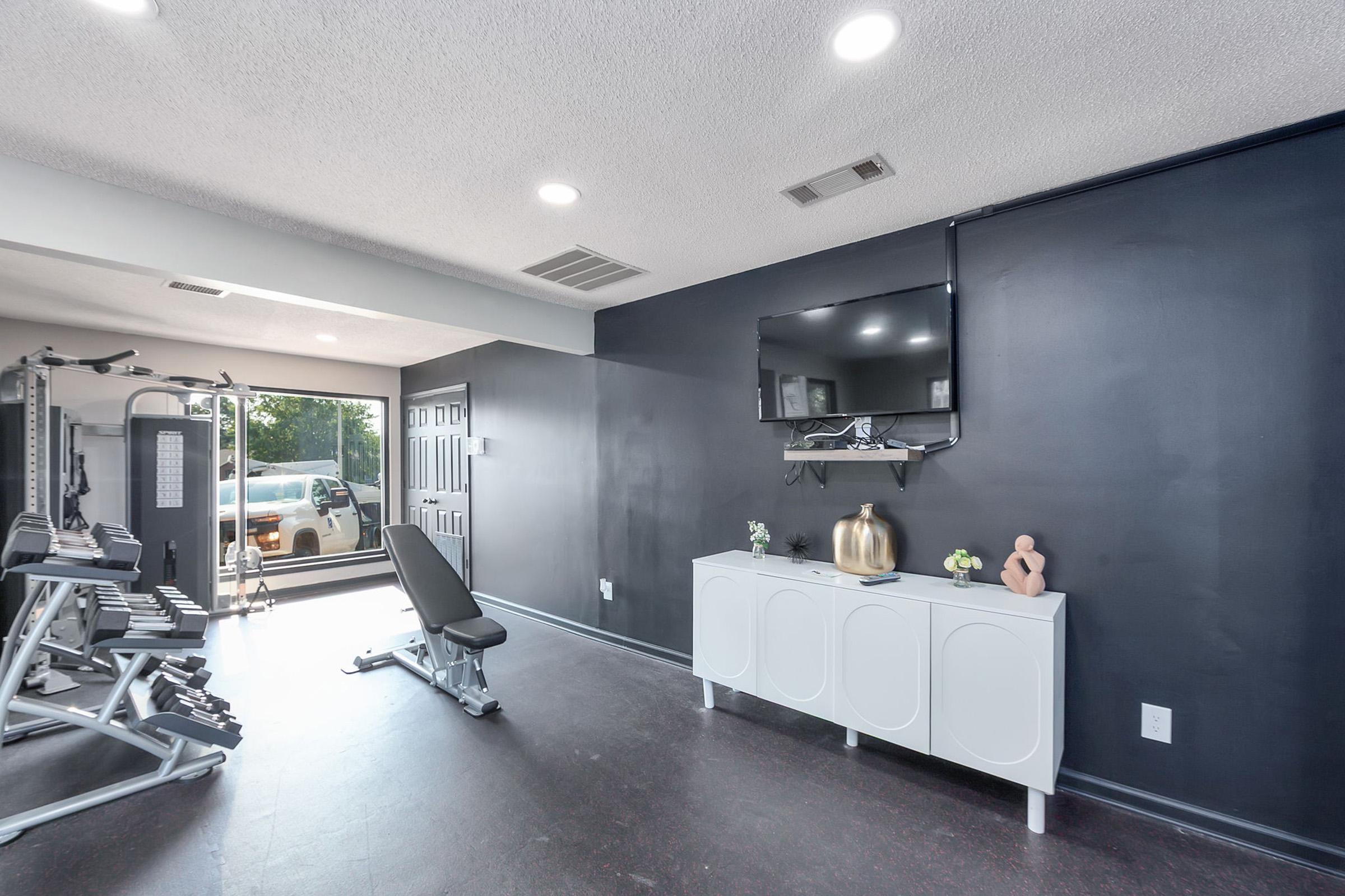 A modern gym space featuring exercise equipment, a weight bench, and a television mounted on a dark wall. Natural light streams in from large windows, highlighting minimalist decor with a white cabinet and decorative items. The room has a clean and contemporary design suitable for workouts.