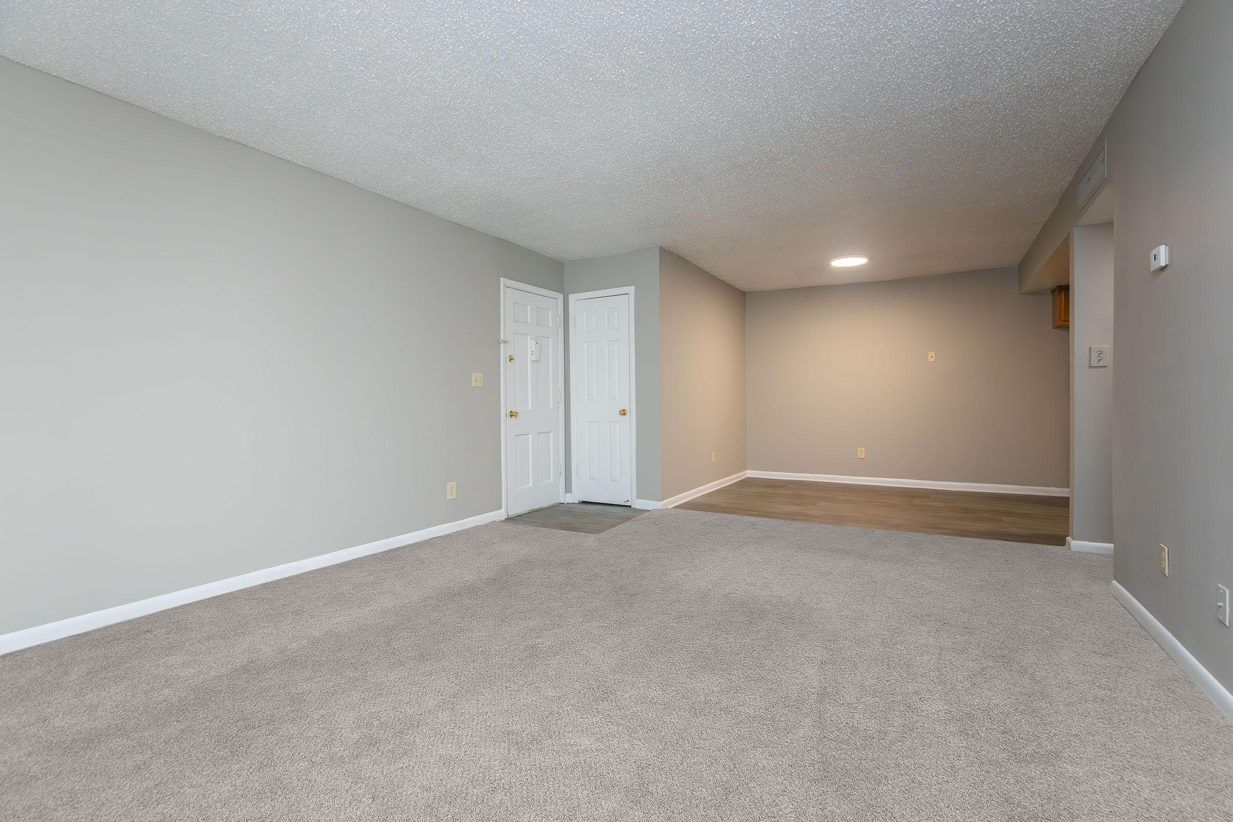 Spacious, empty room with light gray walls and carpeted flooring. Two white doors on the left lead to an entryway, while the right side shows a transition to another room. Soft overhead lighting provides ample illumination, creating a welcoming atmosphere. Ideal for various interior design setups.