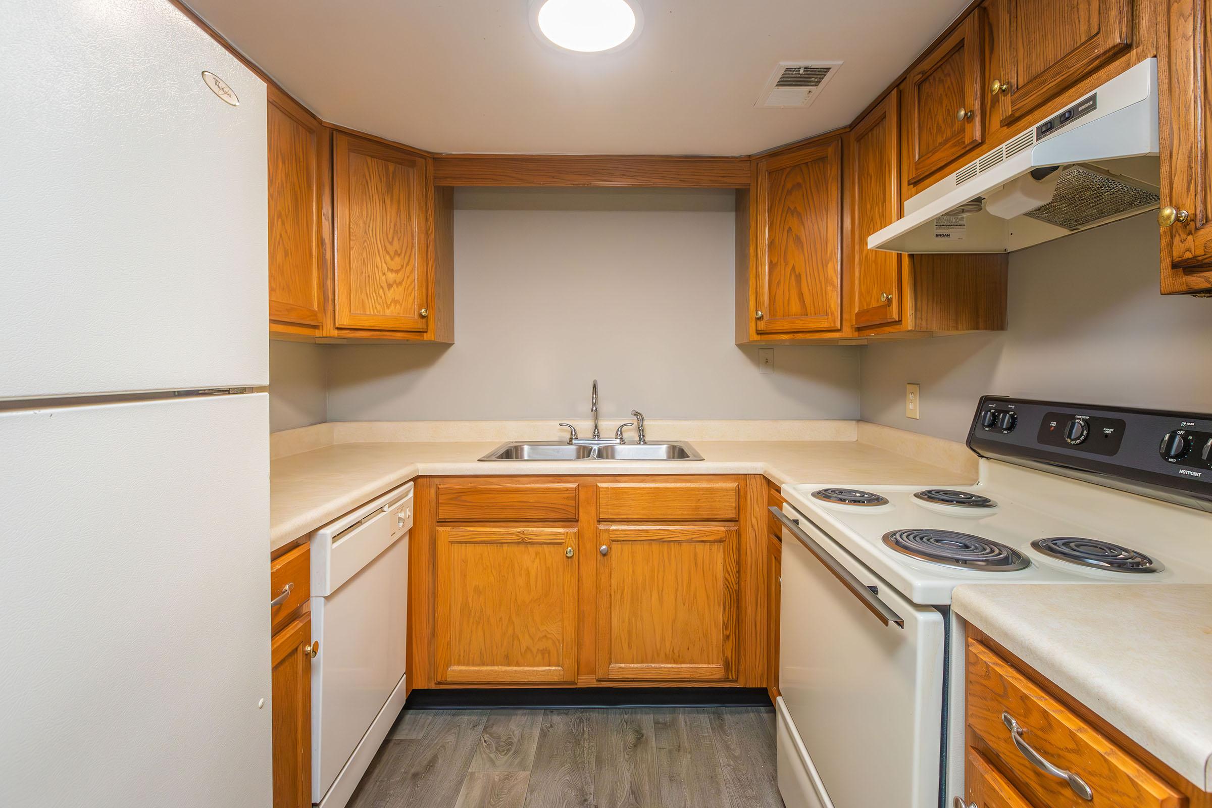 Compact kitchen featuring wooden cabinets, a double sink, and white appliances including a refrigerator, dishwasher, and stove. The space has a neutral-colored wall and a ceiling light, creating a warm and inviting atmosphere. Counter space is available for food preparation.