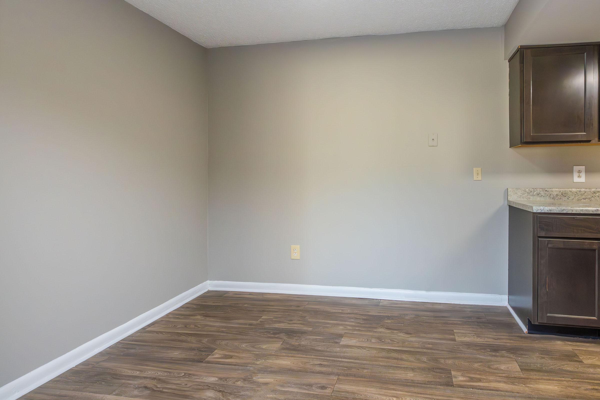 Empty room with light gray walls and a wood-like floor. A small section of a kitchen counter with dark cabinets is visible on the right side of the image. The wall is bare, creating a spacious and minimalistic atmosphere.