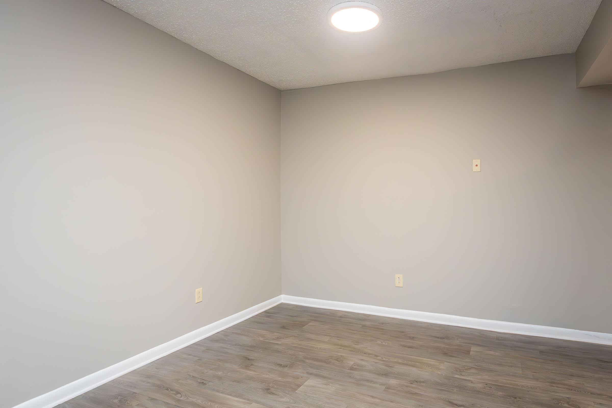 Empty interior room with light gray walls and a smooth wooden floor. A circular ceiling light provides illumination. The space is devoid of furniture or decoration, creating a minimalist and open atmosphere.