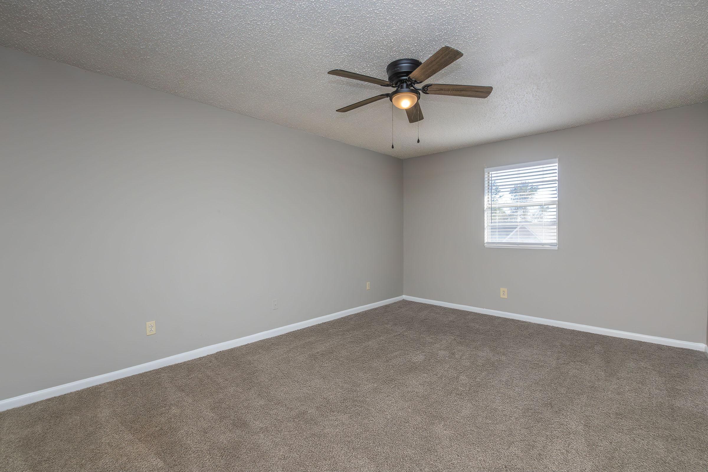 Empty room with light gray painted walls and a ceiling fan. The floor is covered in beige carpet. A window with white blinds allows natural light to enter, illuminating the space. The room has a clean and minimalist appearance, suitable for various uses.