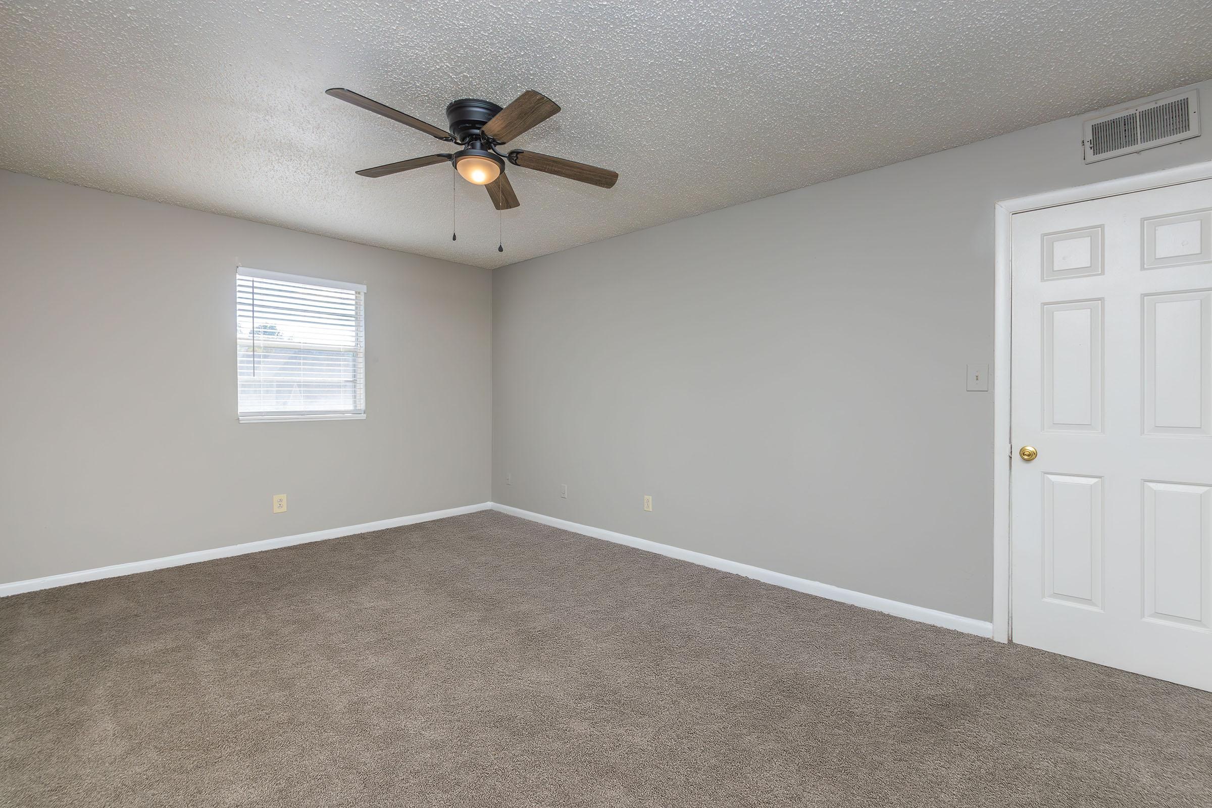 A spacious, empty room with tan carpet, a ceiling fan, and a single window allowing natural light. The walls are painted in a light gray color, and there is a white door on the right side of the image. The overall appearance is clean and ready for personal decoration.