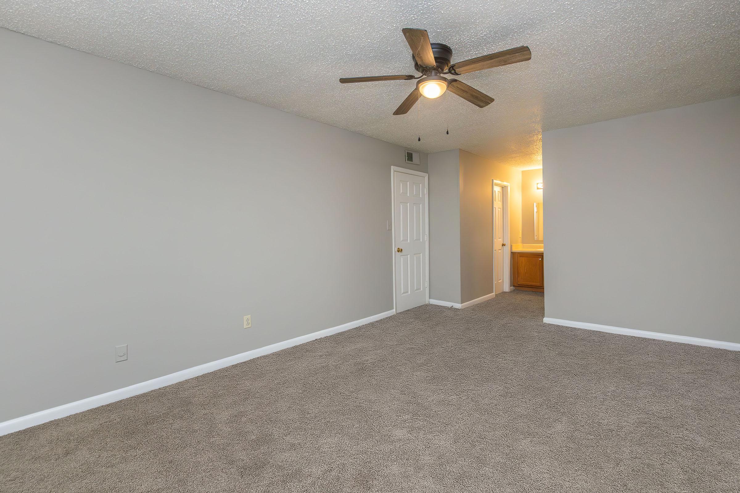 A spacious room with light gray walls and plush beige carpet. The ceiling features a ceiling fan with a light. On the far side, there is a doorway leading to a bathroom area. Natural light comes from an open door, illuminating the room's neutral tones.