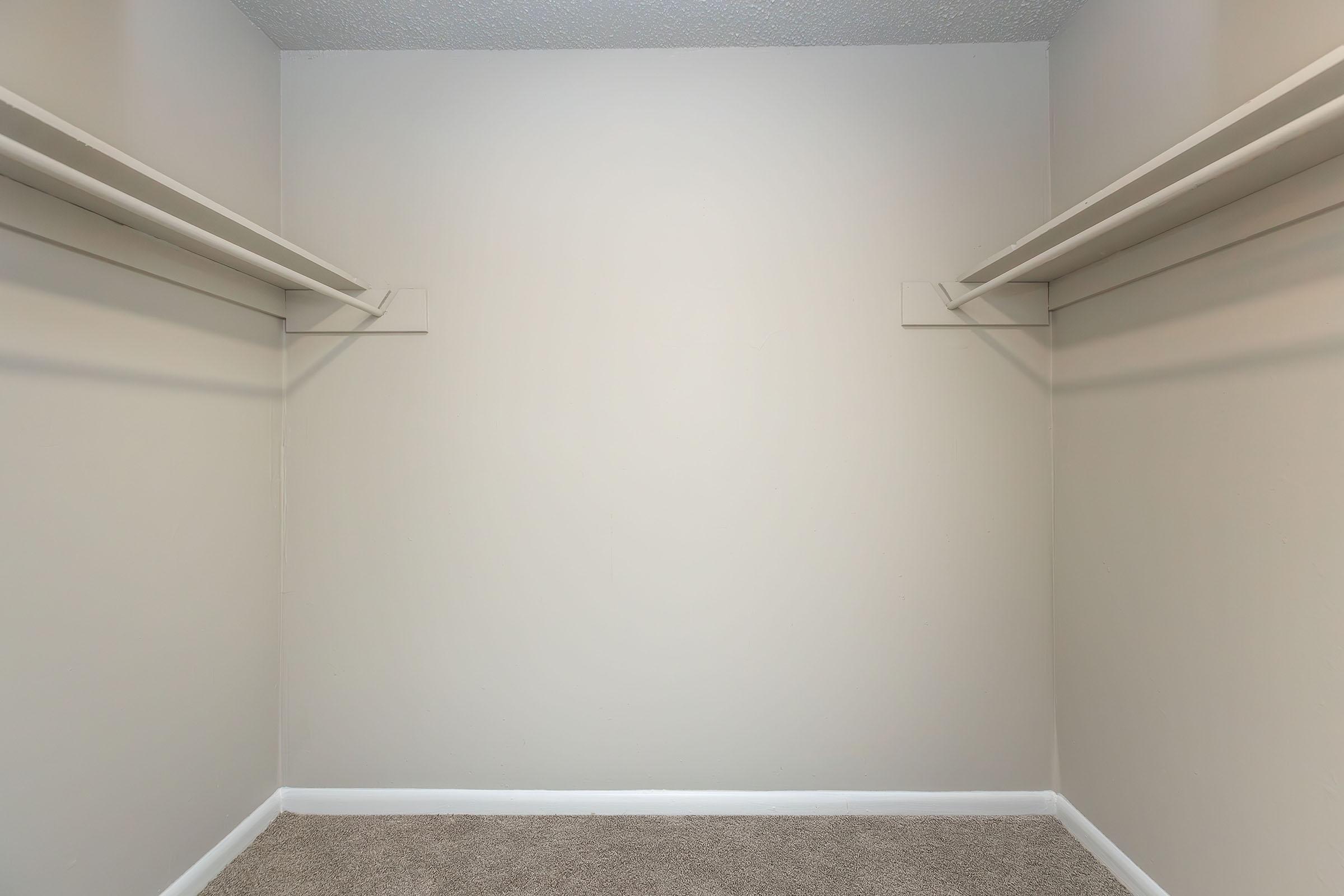 Empty closet with two shelves mounted on the walls. The walls are painted a light beige color, and the floor is covered with tan carpet. There are no items or decorations present, creating a minimalistic and open space.