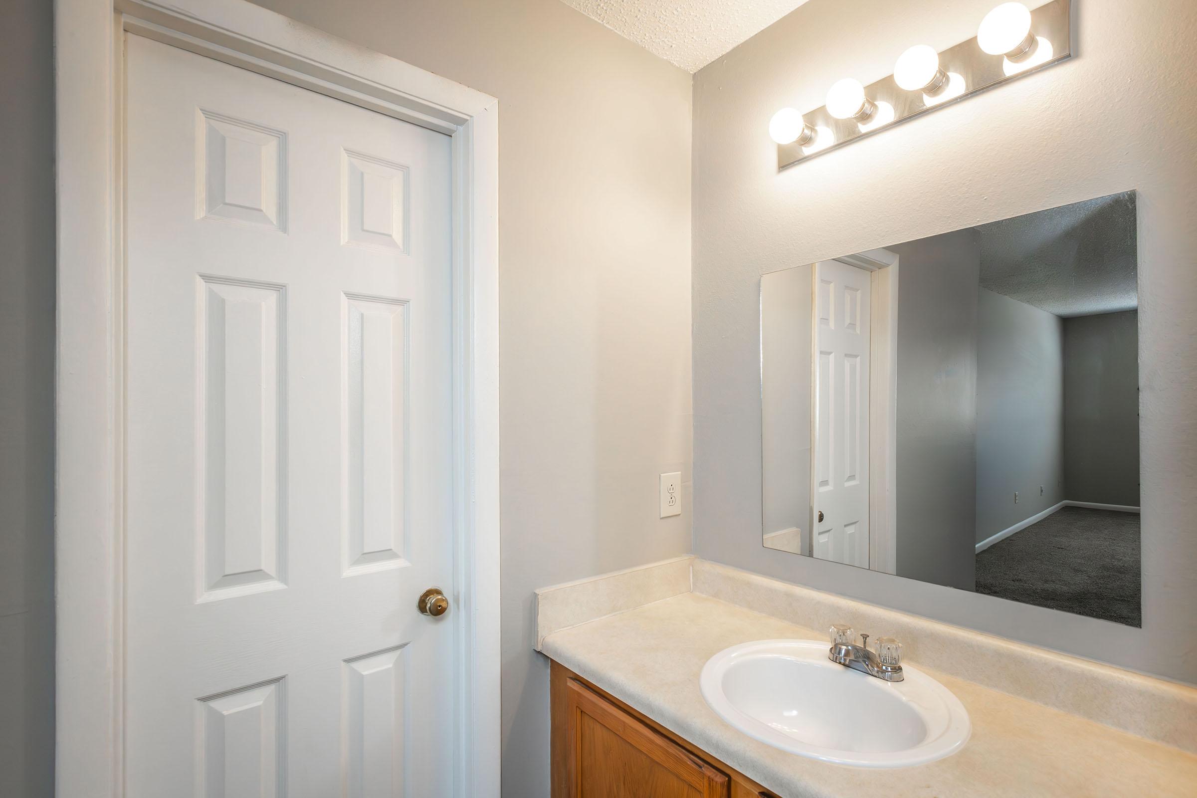 A well-lit bathroom featuring a light fixture above a mirror, a sink with a simple faucet, and a wooden cabinet below. A closed door is visible on the left, while the wall is painted in a soft gray color. The floor appears carpeted in a neutral tone, adding a cozy touch to the space.