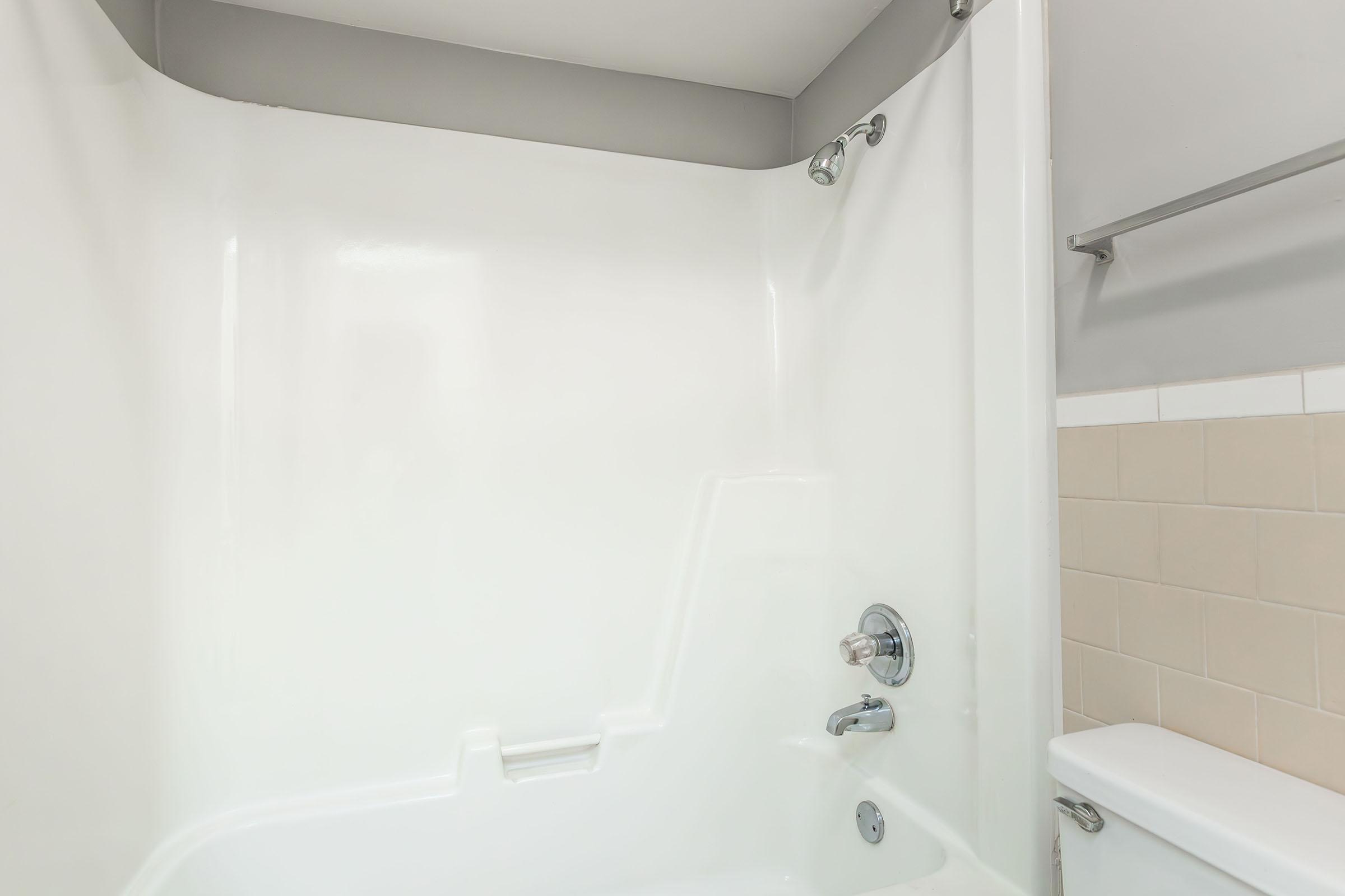 A clean, bright bathroom featuring a white bathtub with a shower head and curtain rod. Light beige tiles line the wall next to the bathtub, and a toilet is visible in the corner. The overall design is modern and minimalistic, emphasizing a neat and tidy appearance.