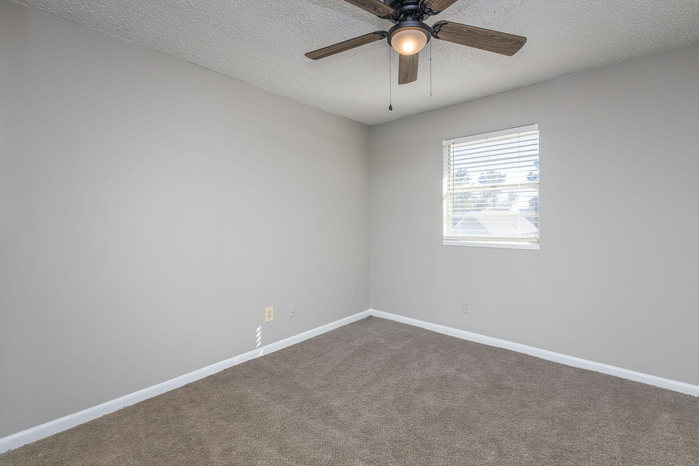 A small, empty bedroom featuring light gray walls, a ceiling fan with wooden blades, and a window with blinds. The floor is covered in beige carpet, and there are no furnishings in the room, providing a clean, spacious appearance.